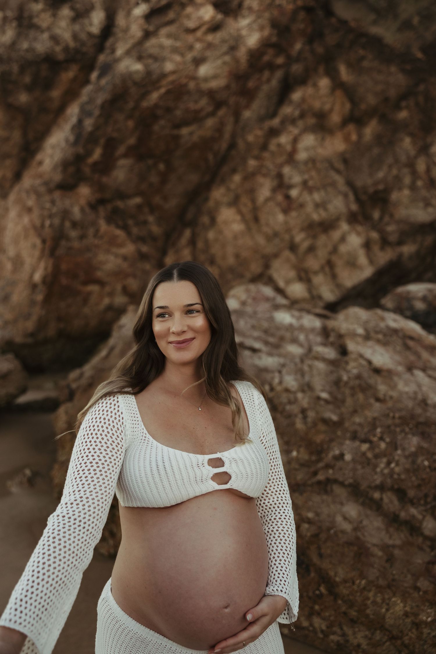 Pregnant woman in white crochet top poses against rocky backdrop during maternity photoshoot.