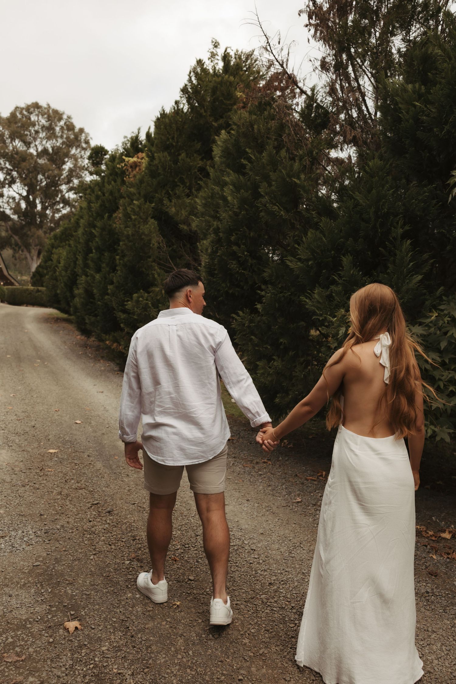 A couple in white outfits walking hand in hand down a dirt path surrounded by evergreen trees.