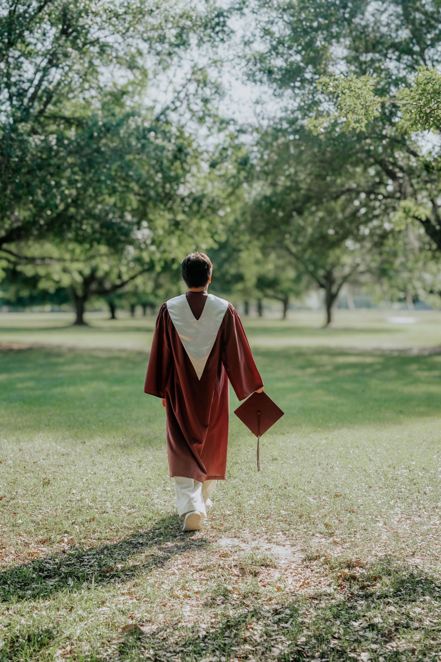 A graduate in maroon cap and gown walks through a lush green park, holding their mortarboard, viewed from behind.