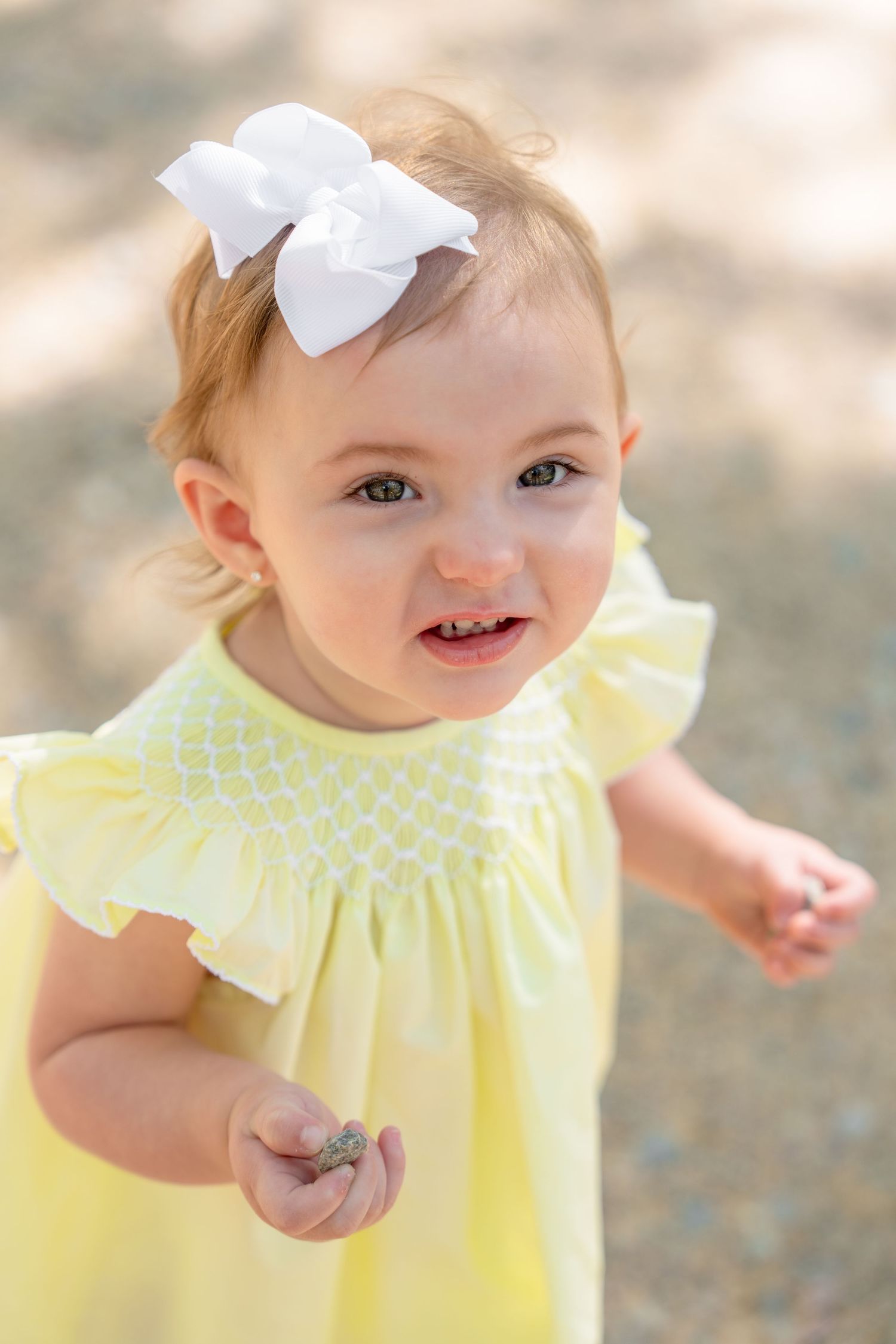 Toddler girl in yellow smocked dress with white bow, holding a small object outdoors.