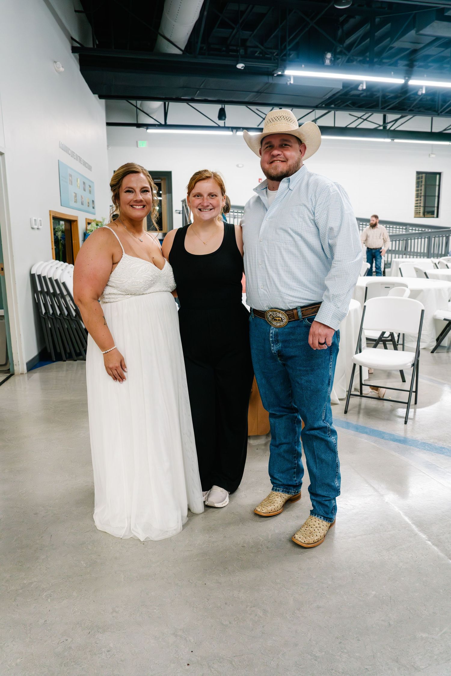 Three people posing together at a wedding reception, bride in white dress, guest in black, man in cowboy hat and jeans.