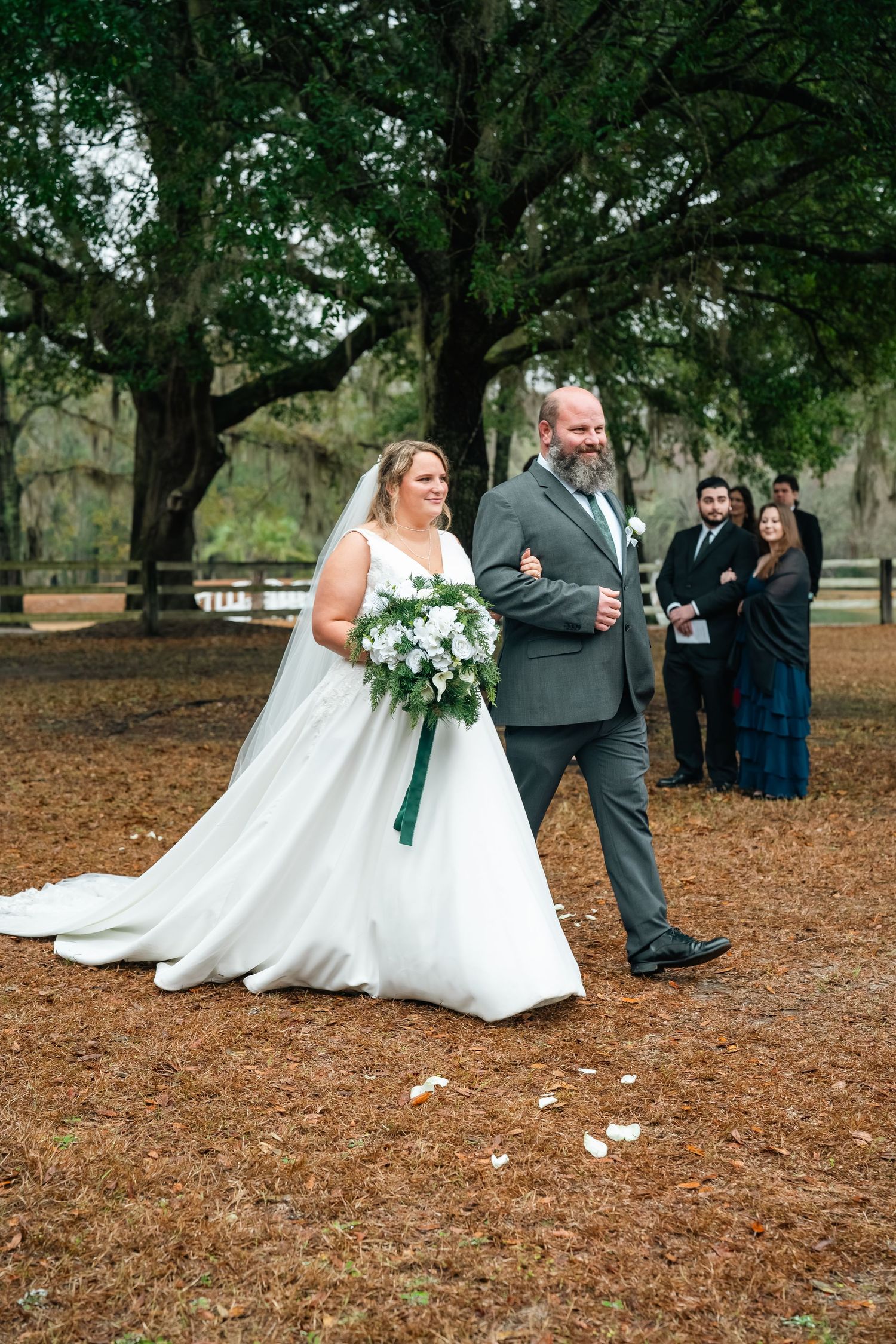 Bride in white gown holding bouquet walks with bearded groom in gray suit at outdoor wedding ceremony under oak trees.
