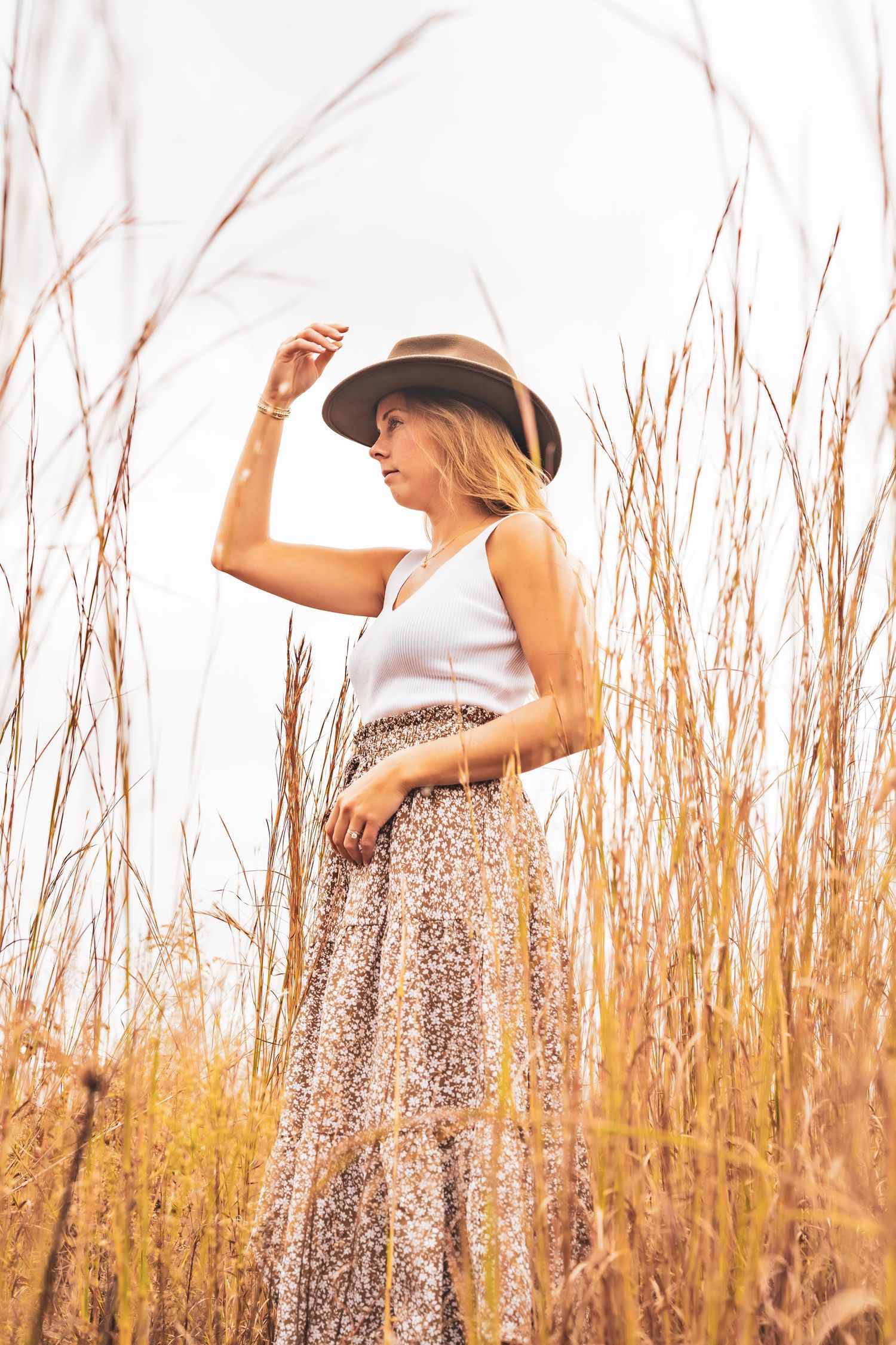 Sunlit portrait series among golden dried grass wearing a brown hat and white top.