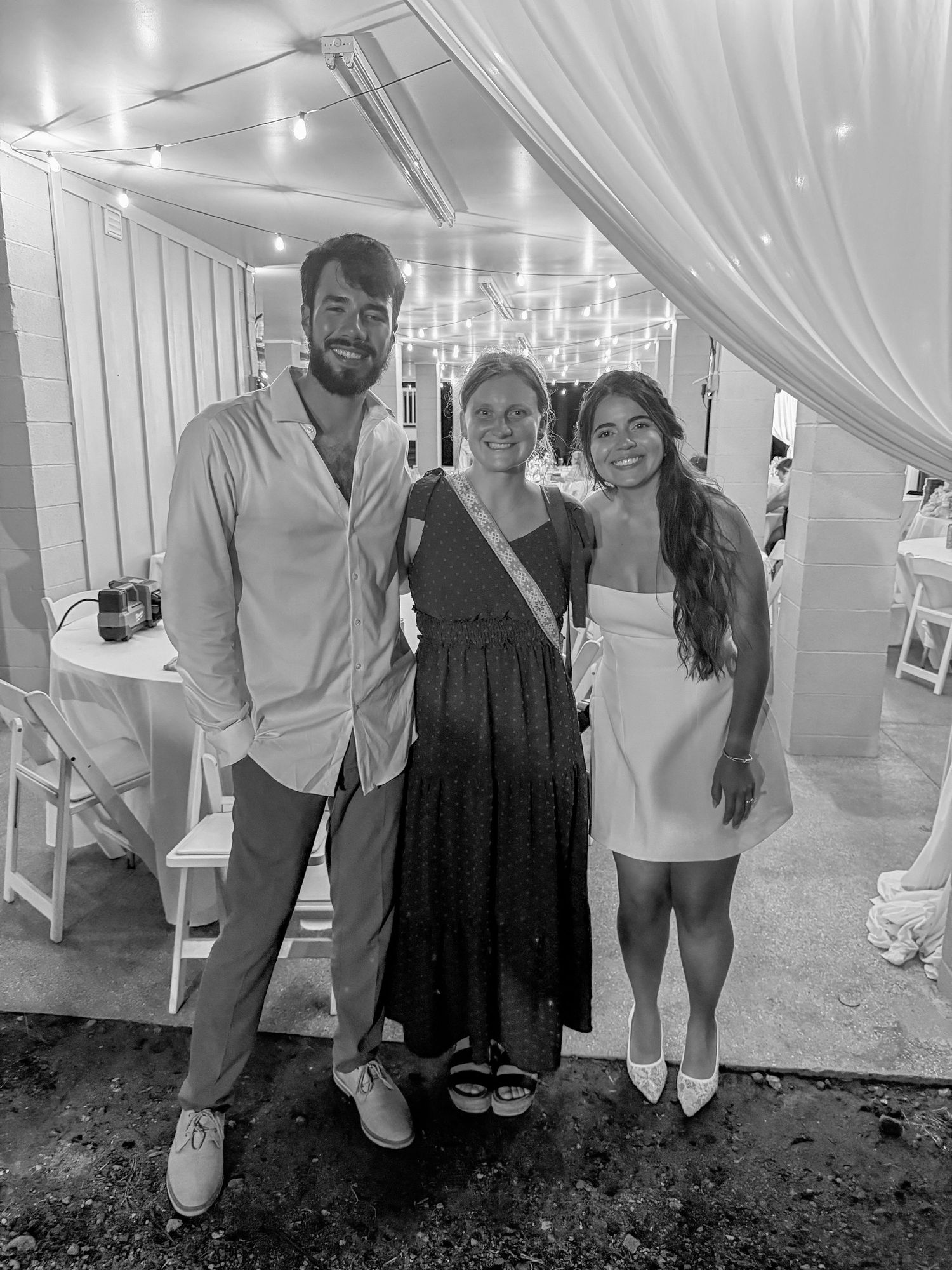 Three people posing together at an elegant outdoor wedding reception under string lights in black and white photo.