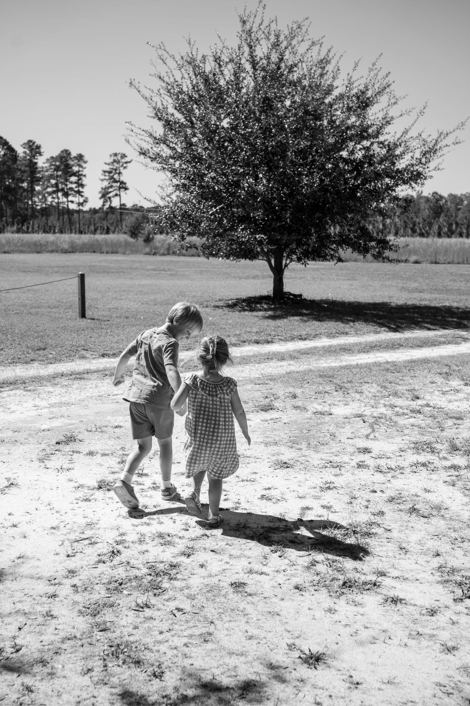 Two figures walking together on a dirt path under a lone tree in black and white.
