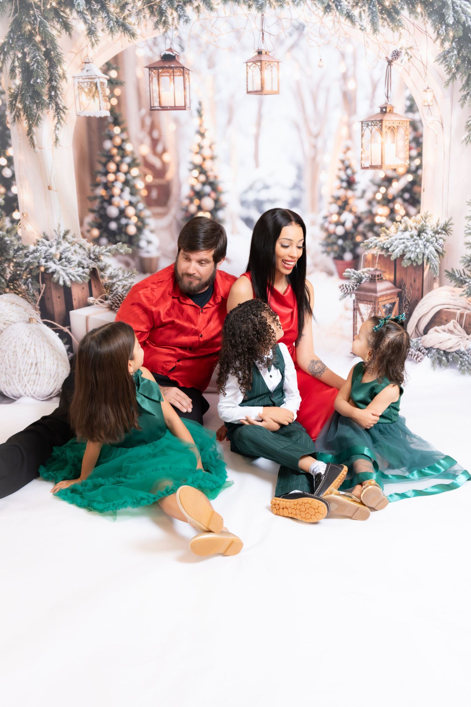 A couple in red and green holiday outfits poses playfully in a winter wonderland photo shoot setting with Christmas decor.