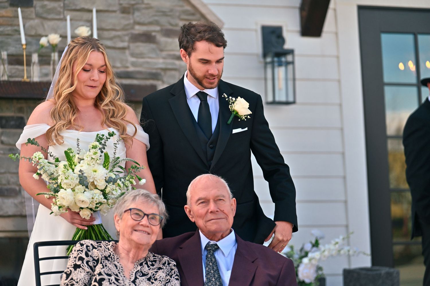 Wedding couple in formal attire poses with seated elderly guests outside a white farmhouse, bride holding white floral bouquet.