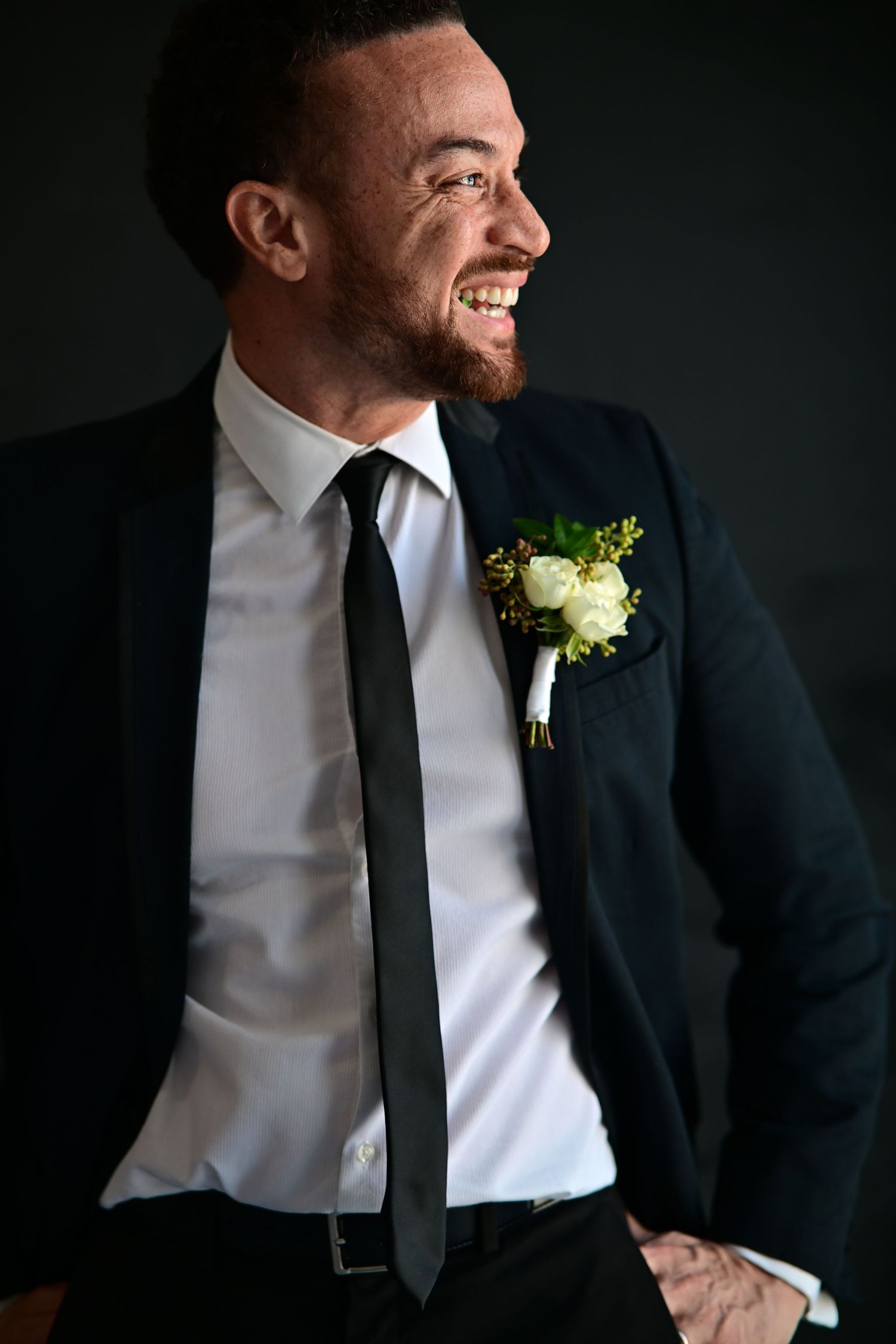 Groom in classic black suit and tie with white boutonniere laughs joyfully against dark background.