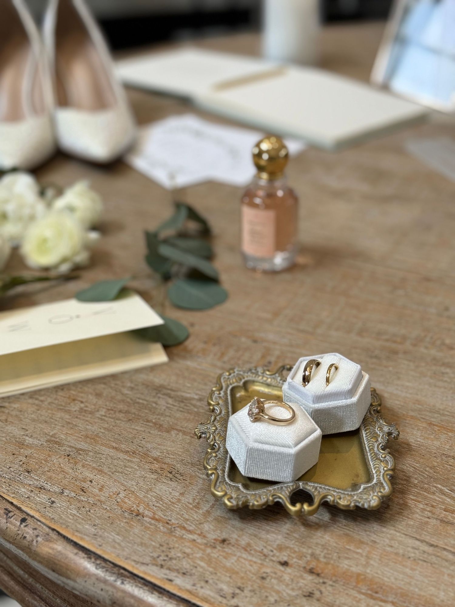 Gold wedding rings displayed in a white velvet box on a vintage tray, with bridal accessories on a wooden table.