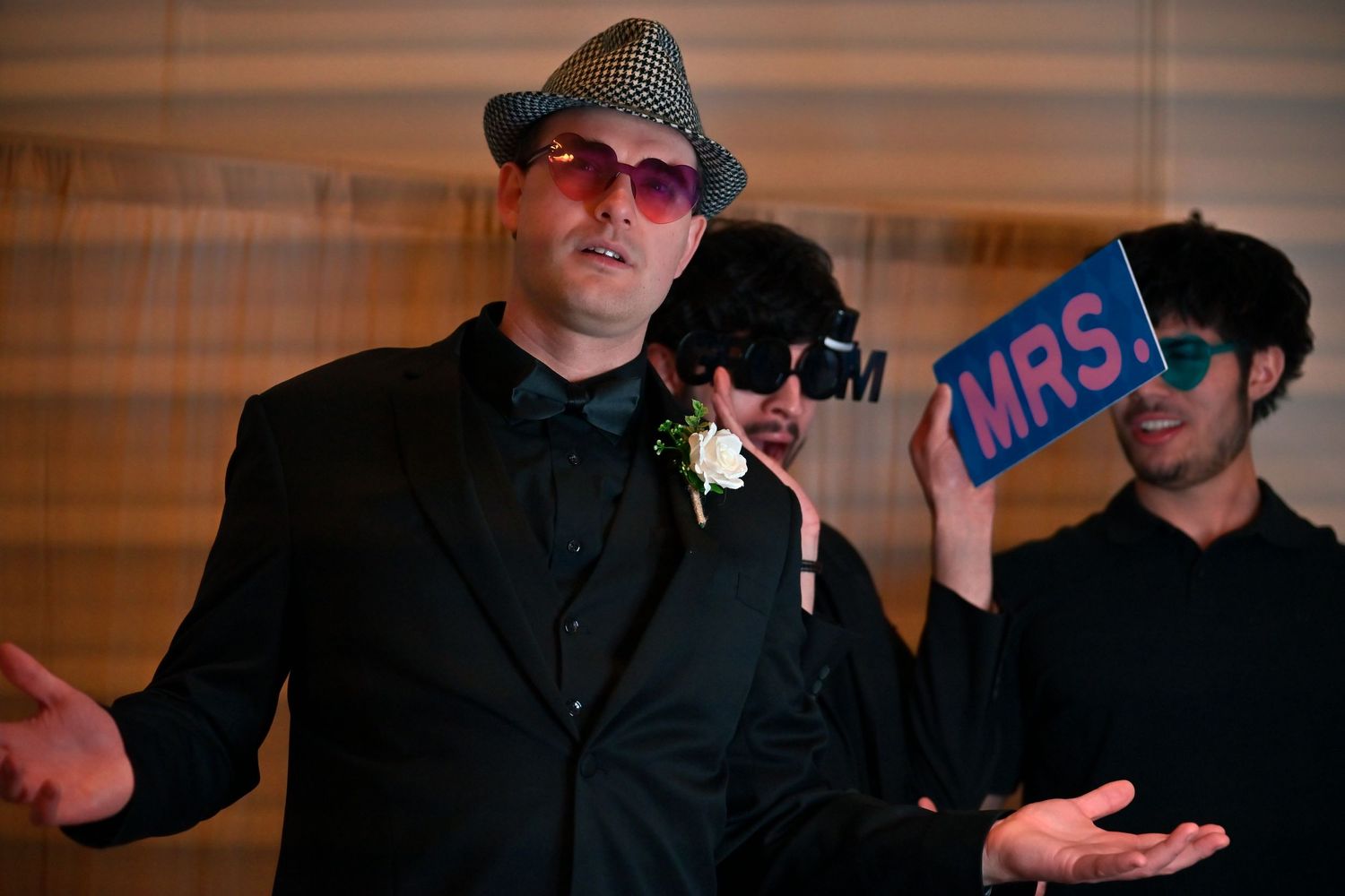 Men in suits and hats at a wedding reception, one holding a blue MRS. sign, posing playfully on stage.