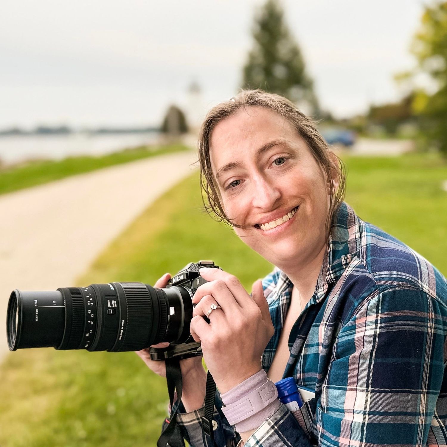 A smiling woman holding a DSLR camera with a zoom lens outdoors near a scenic waterway path.