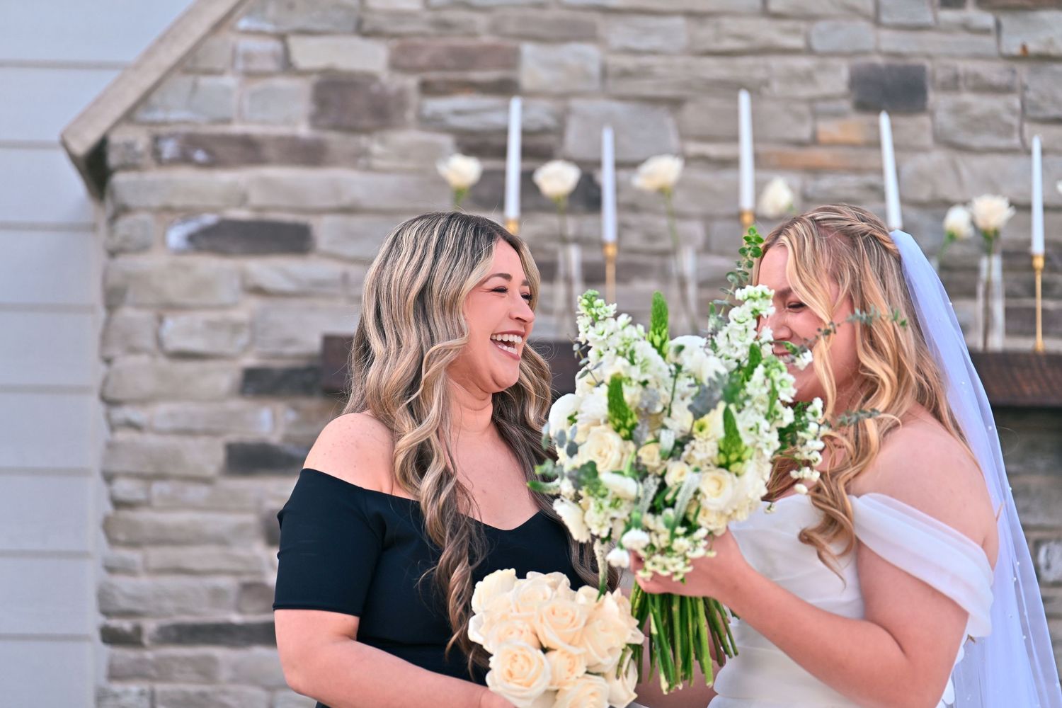 Bride in white gown and veil laughing with bridesmaid in black dress, both holding white floral bouquets outdoors.