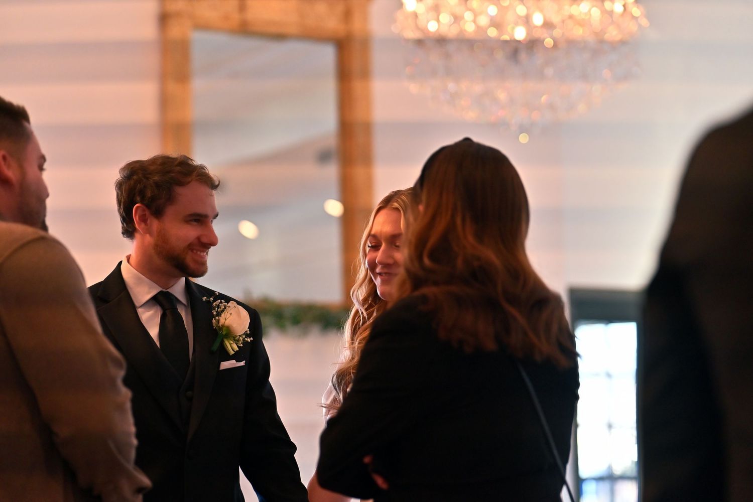 Groom in black suit with boutonniere smiling at guests during elegant wedding reception with chandelier.