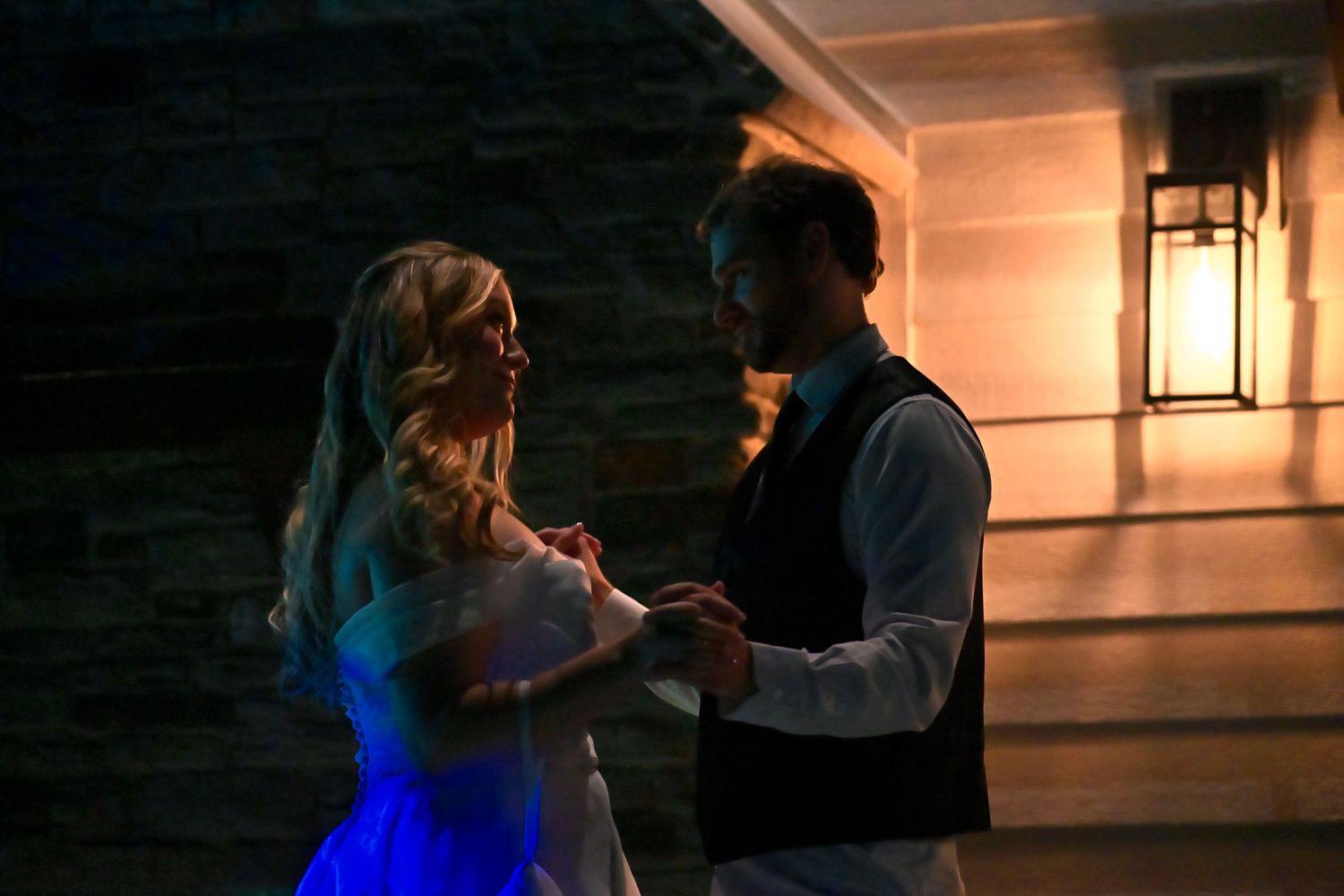 Couple sharing romantic first dance outdoors at night, illuminated by warm wall lantern light.