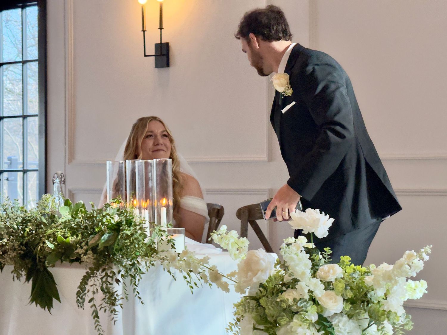 Bride and groom at wedding reception head table with white floral arrangements and greenery.