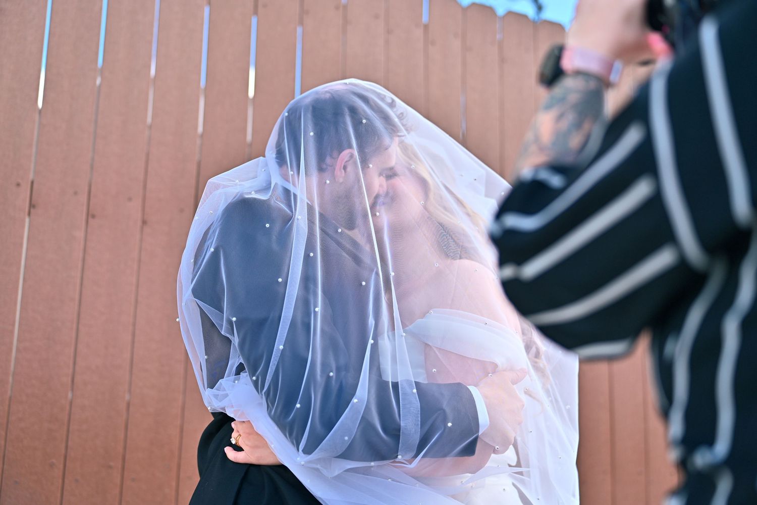 Bride and groom sharing a kiss under a white wedding veil outdoors, with a photographer capturing the moment.