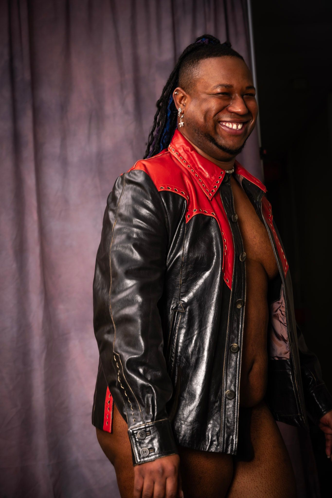 A wrestler in a black and red leather jacket smiles broadly while posing against a gray backdrop.