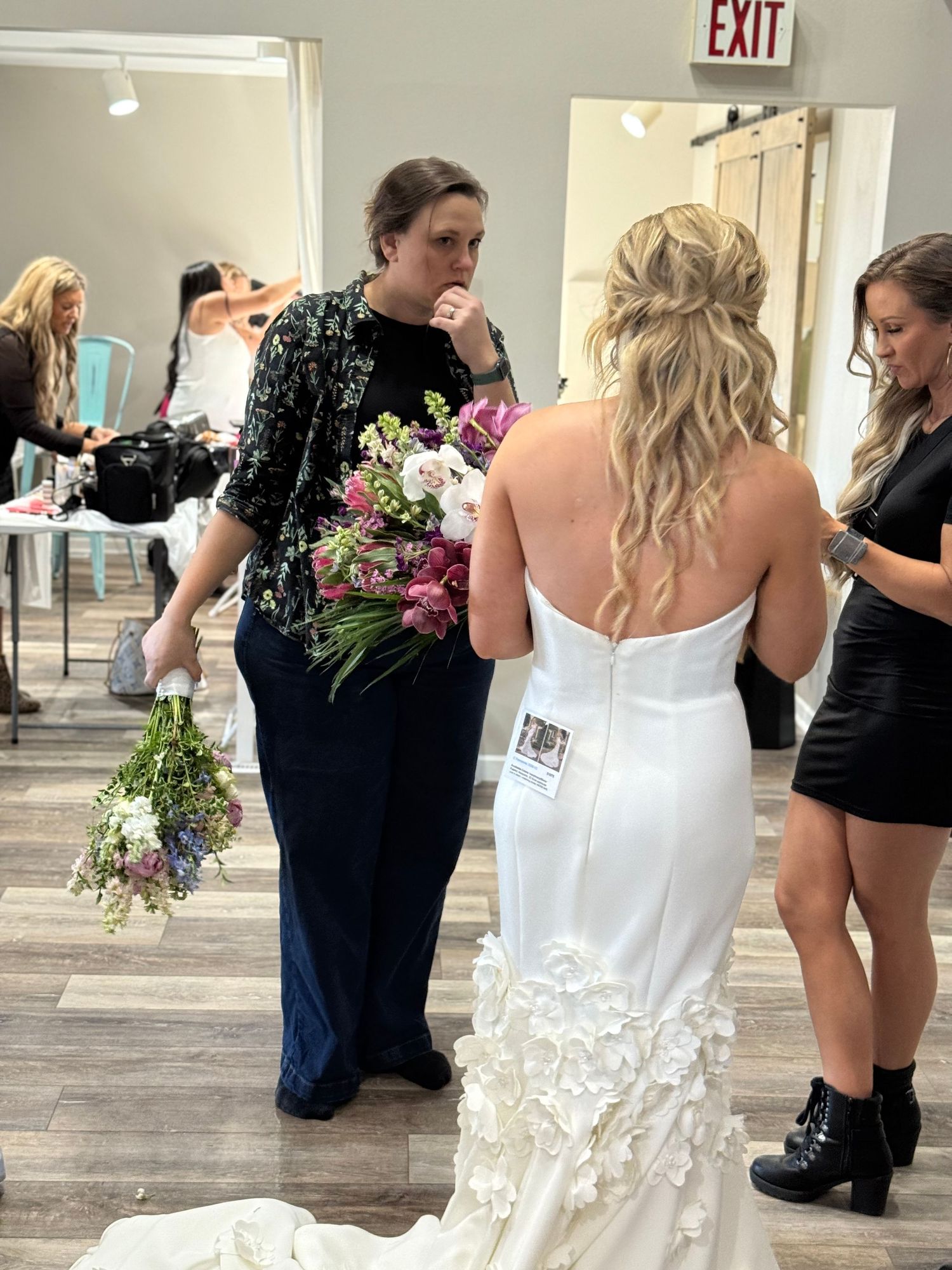 A bride in a white lace-detailed wedding dress stands with others holding floral bouquets in an indoor venue.