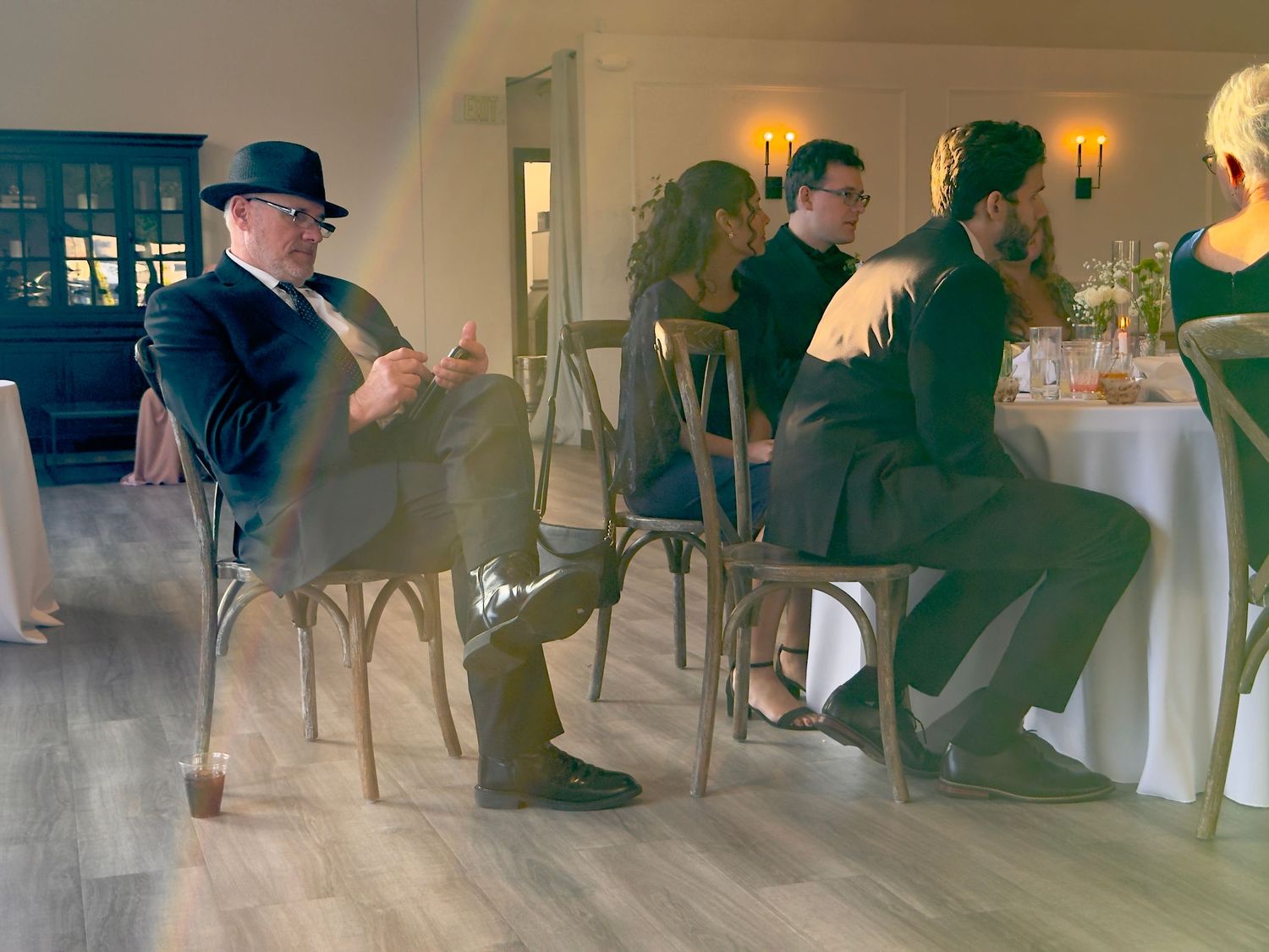 Elegantly dressed guests seated on wooden chairs at an indoor wedding reception venue with warm lighting.