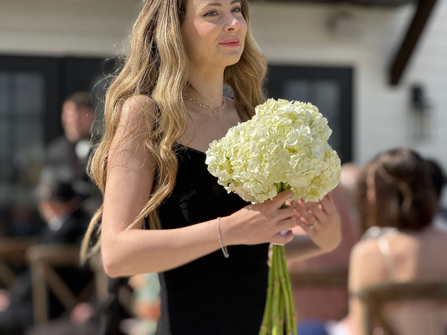 Young woman in black dress holding white hydrangea bouquet at outdoor wedding ceremony.