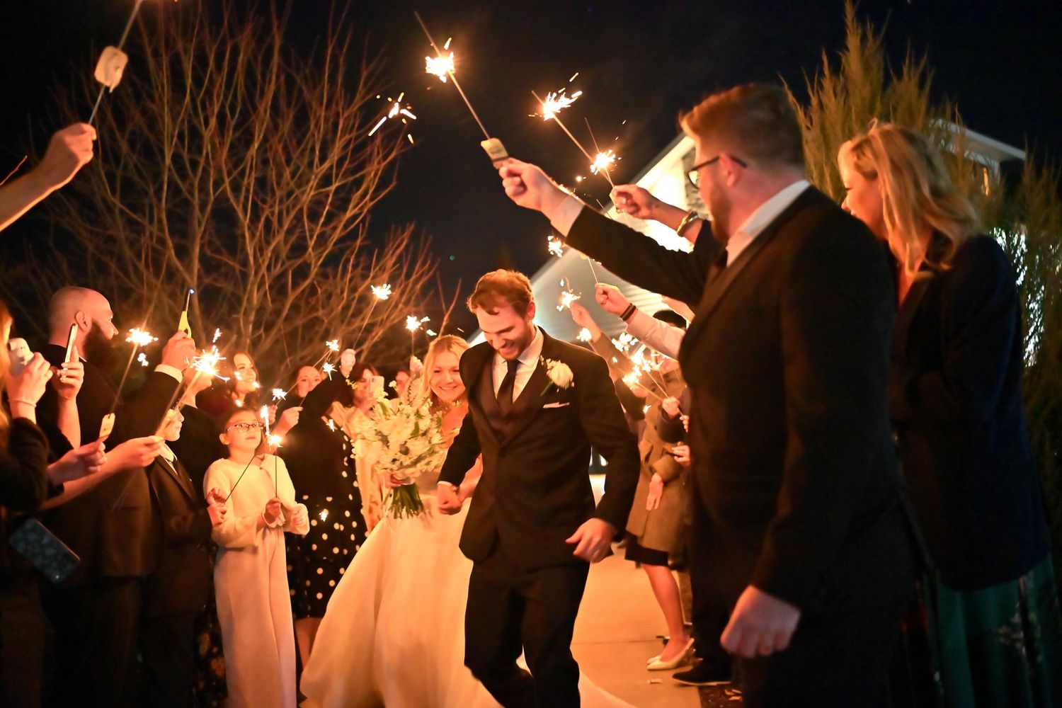 Newlywed couple exits wedding reception through sparkler tunnel at night surrounded by cheering guests.