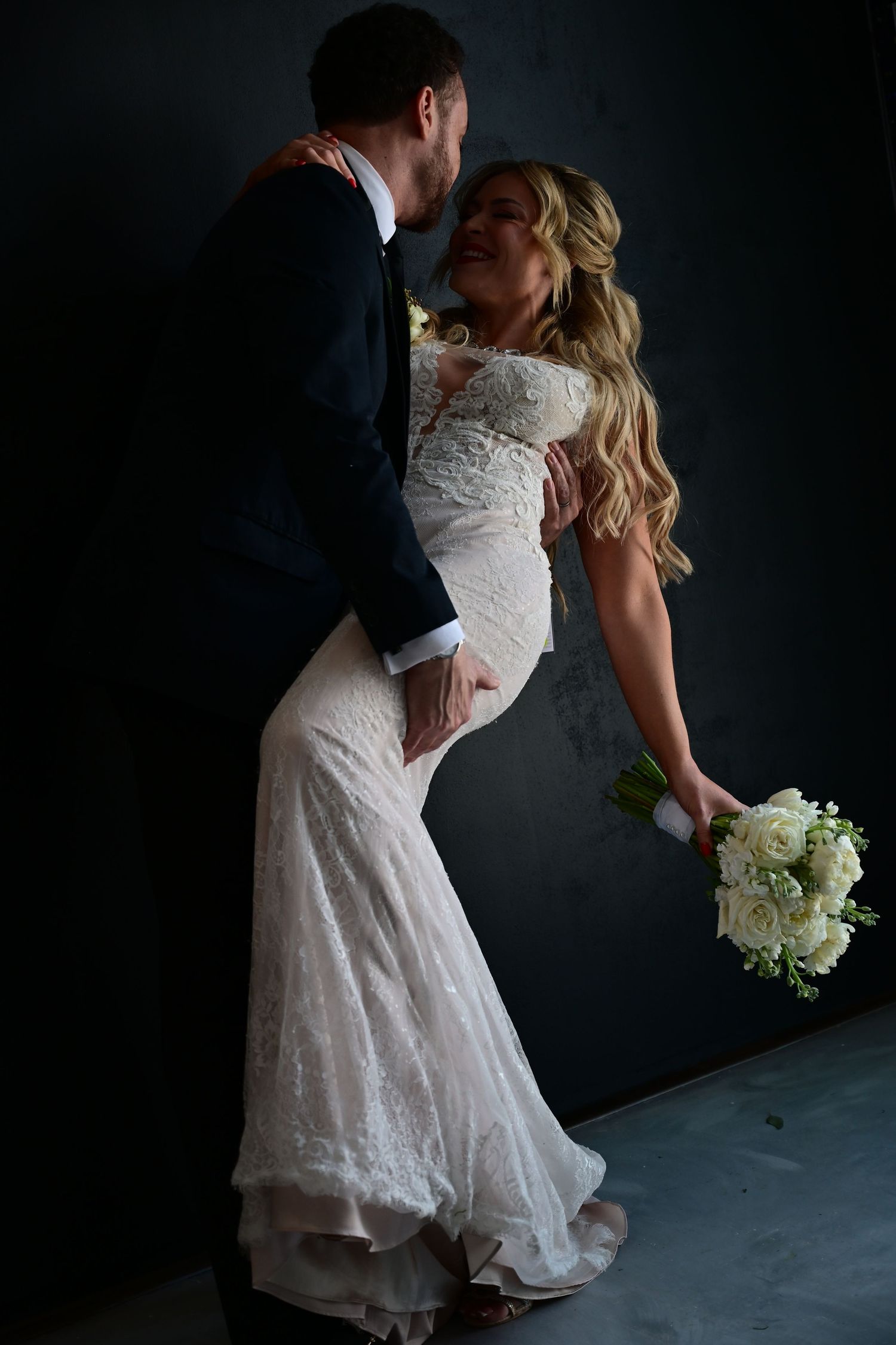 A romantic wedding couple embraces in dramatic lighting against a dark background while holding a white floral bouquet.