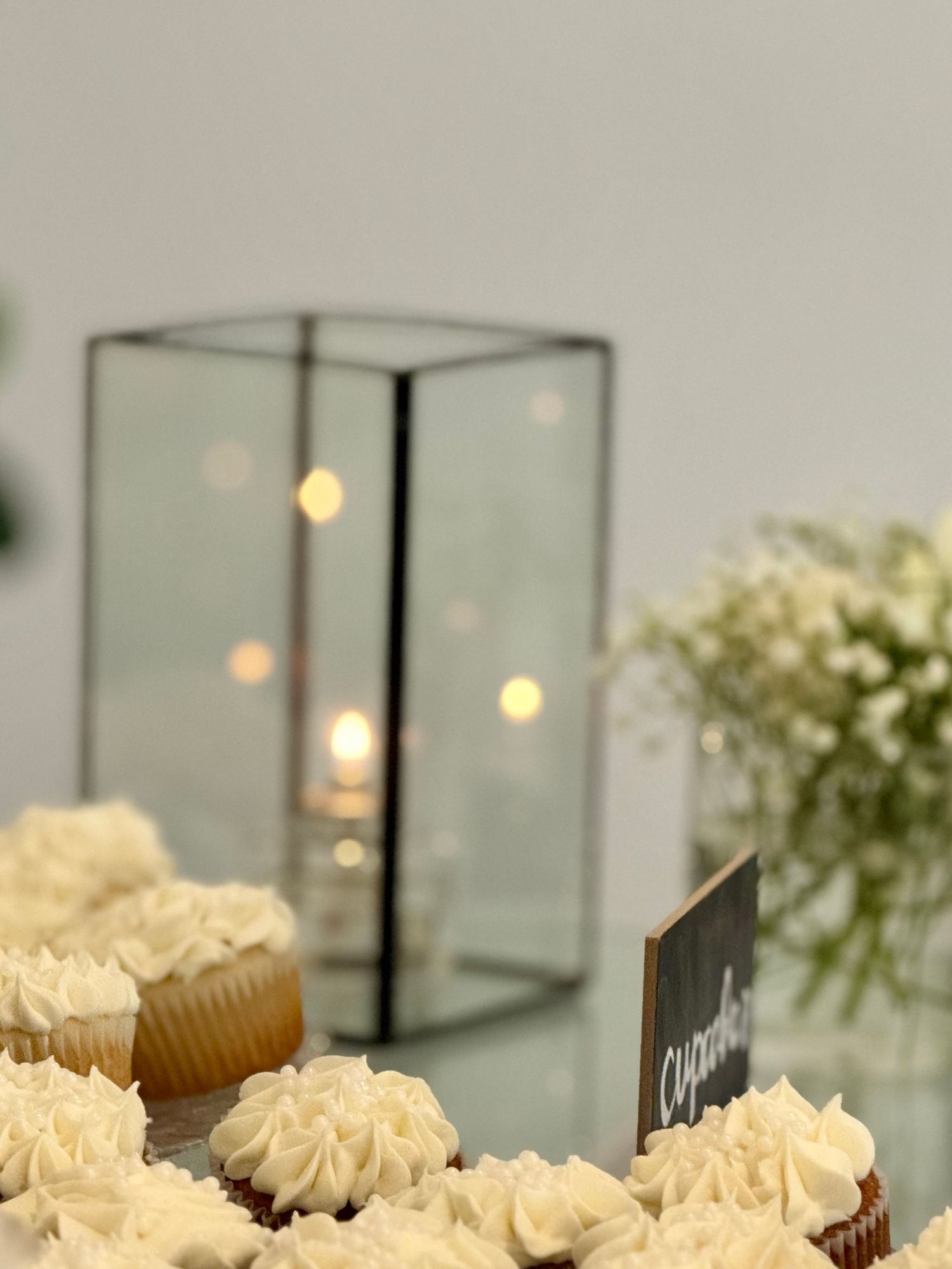 Elegant cupcakes with white frosting displayed on a table with a glass lantern and floral arrangement in the background.