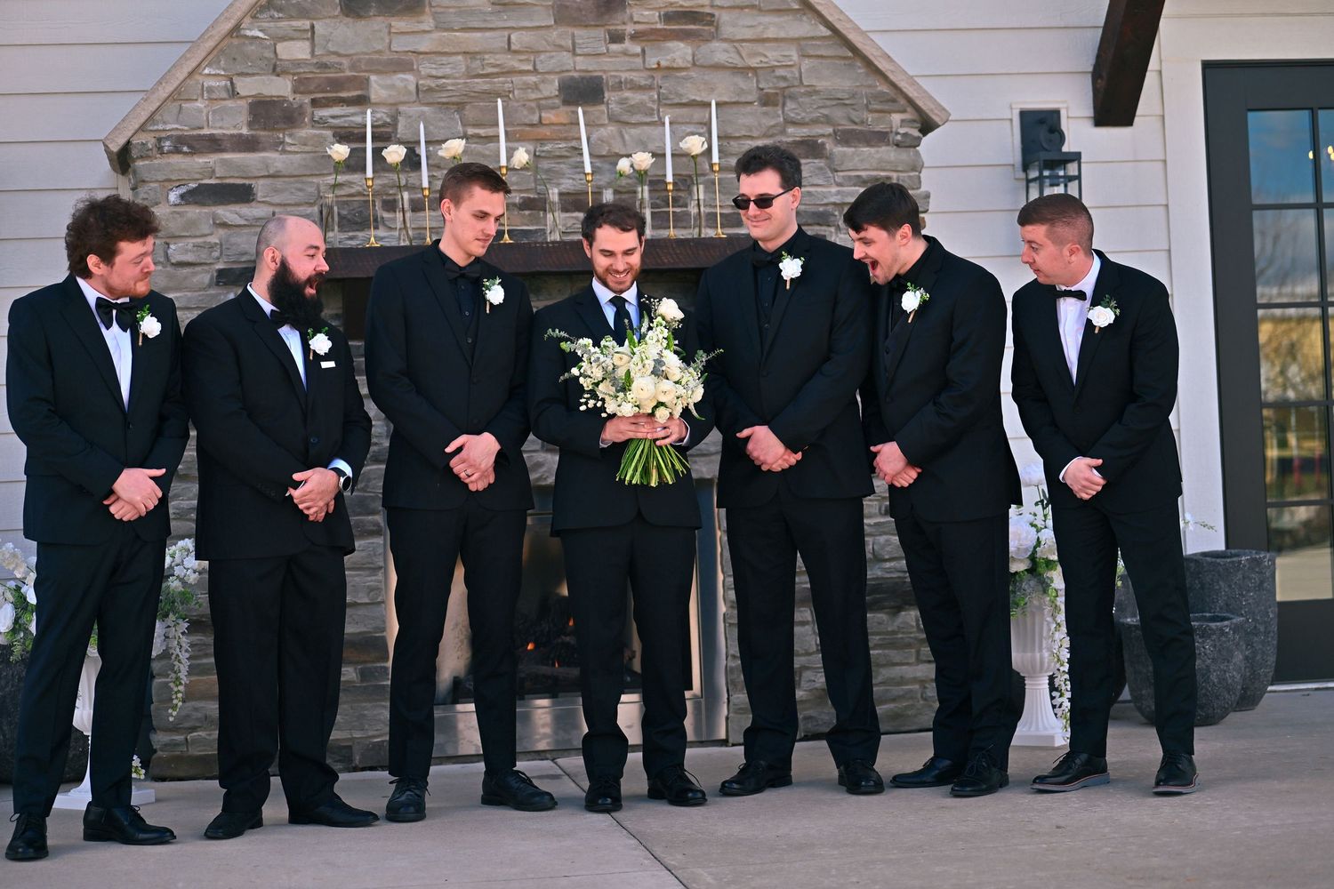 Groom and groomsmen in black tuxedos laughing together outdoors near stone fireplace holding white wedding bouquet.