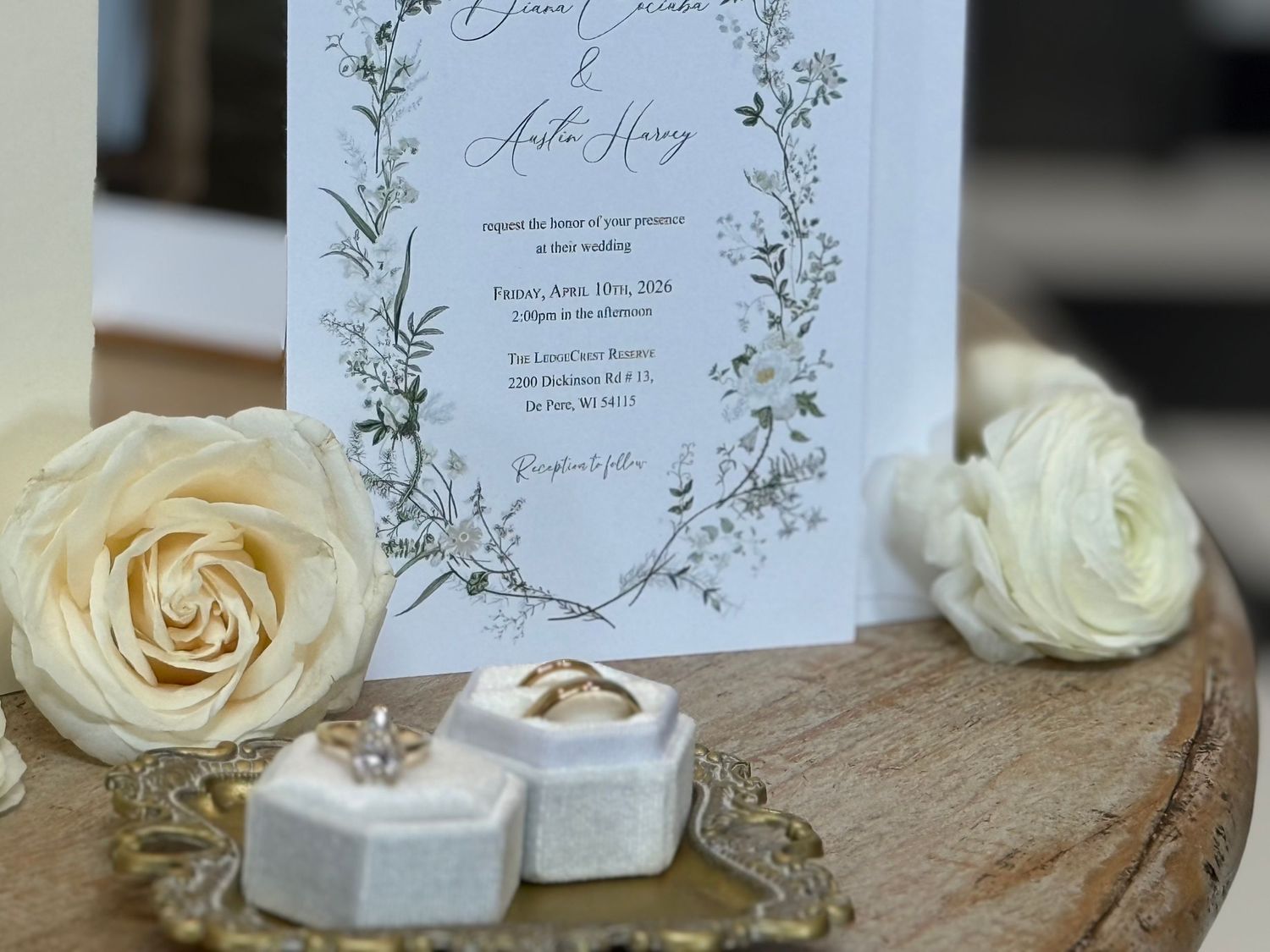 Elegant wedding invitation with floral wreath design, white roses, and diamond ring boxes on a wooden surface.