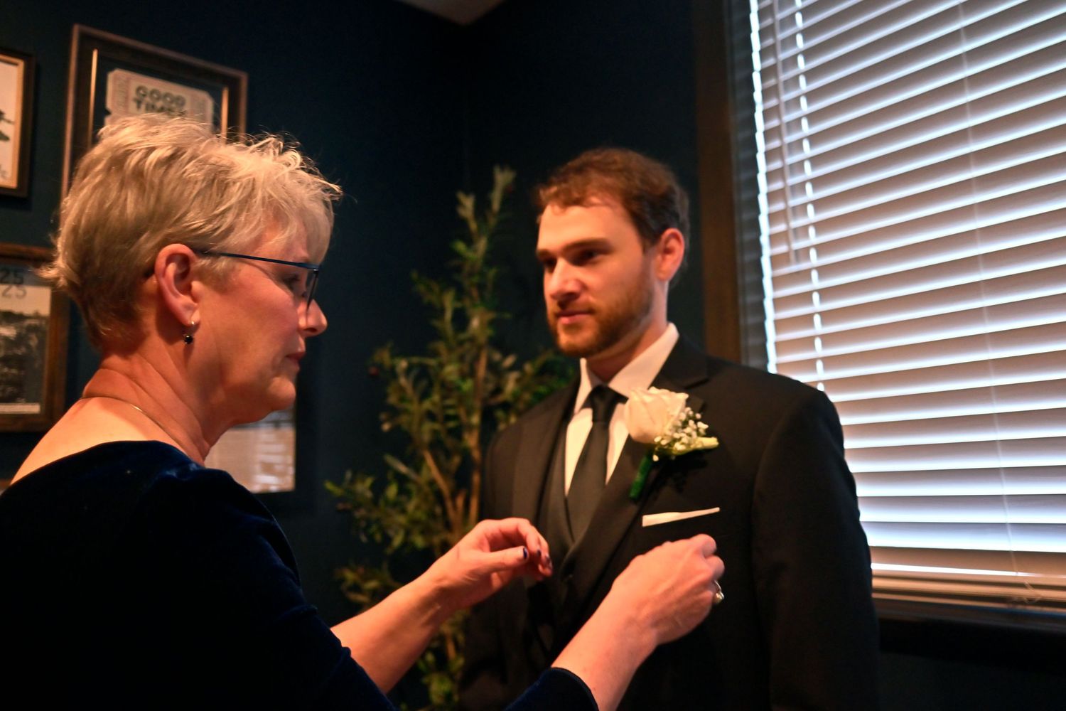 Older woman pinning a boutonniere on a young man in a black suit and tie at a formal event.