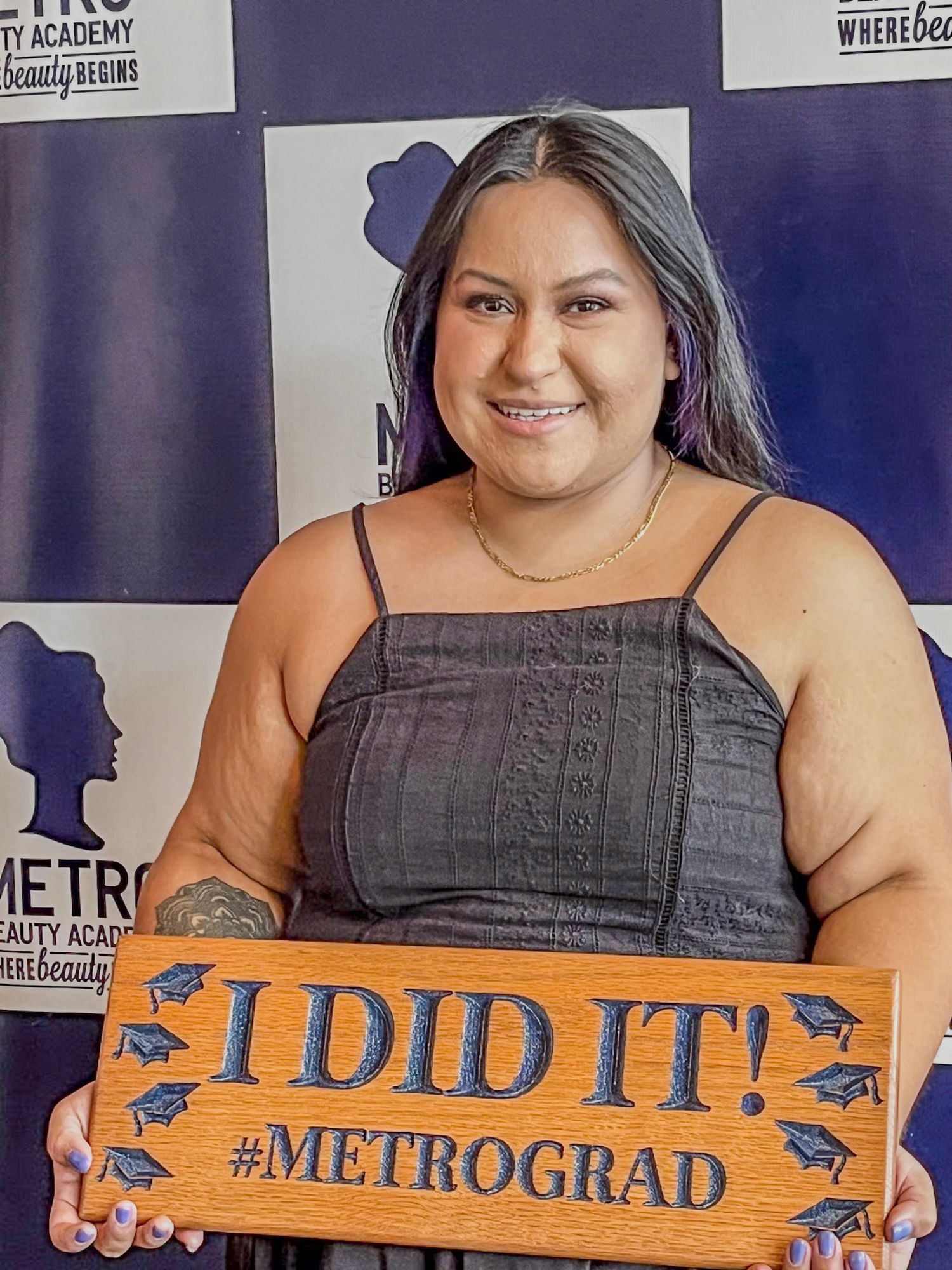 A graduate poses with a wooden 'I DID IT!' sign at Metro Academy against a blue backdrop.