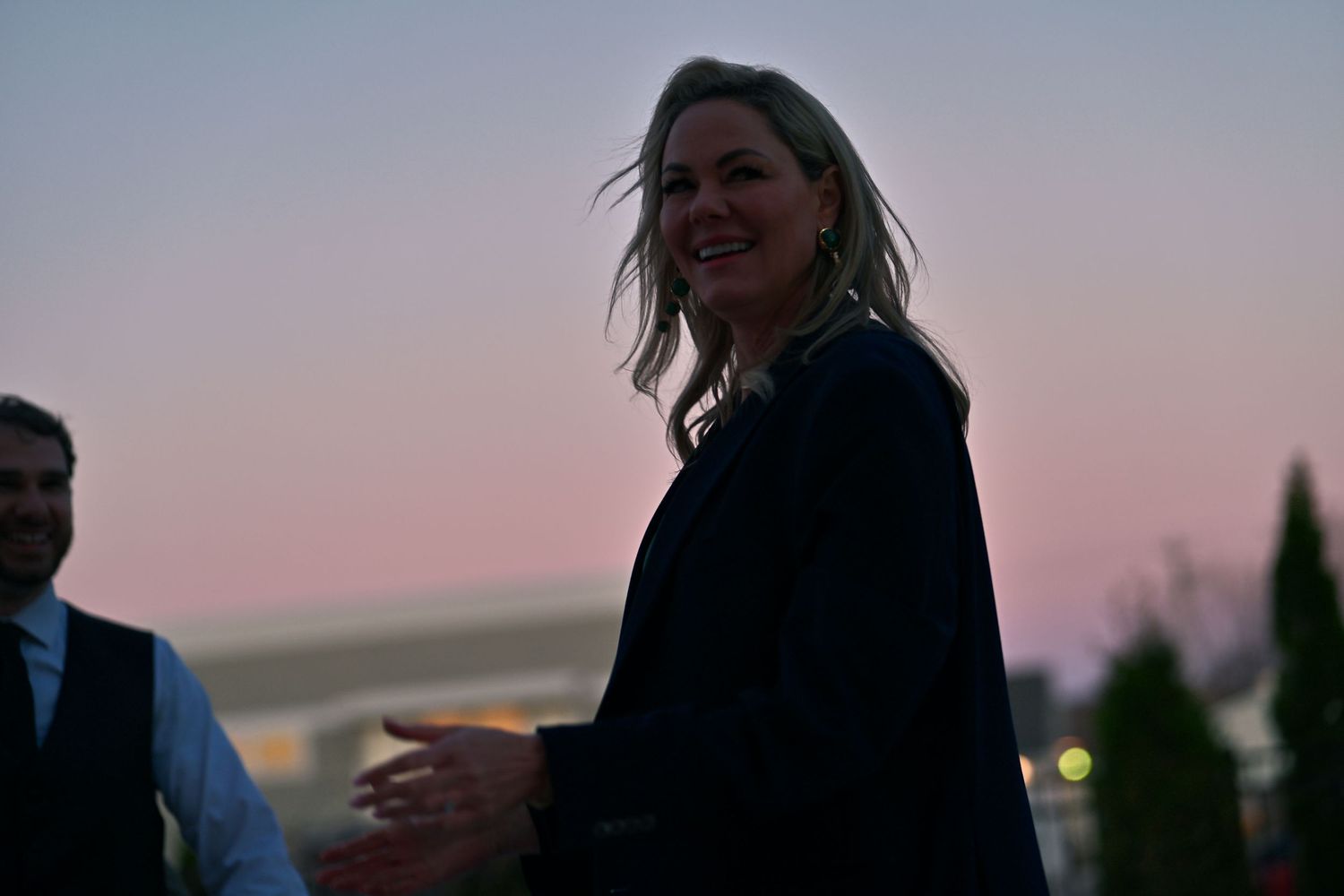 Woman smiling outdoors at dusk with pink sky backdrop, wearing dark blazer at evening event.