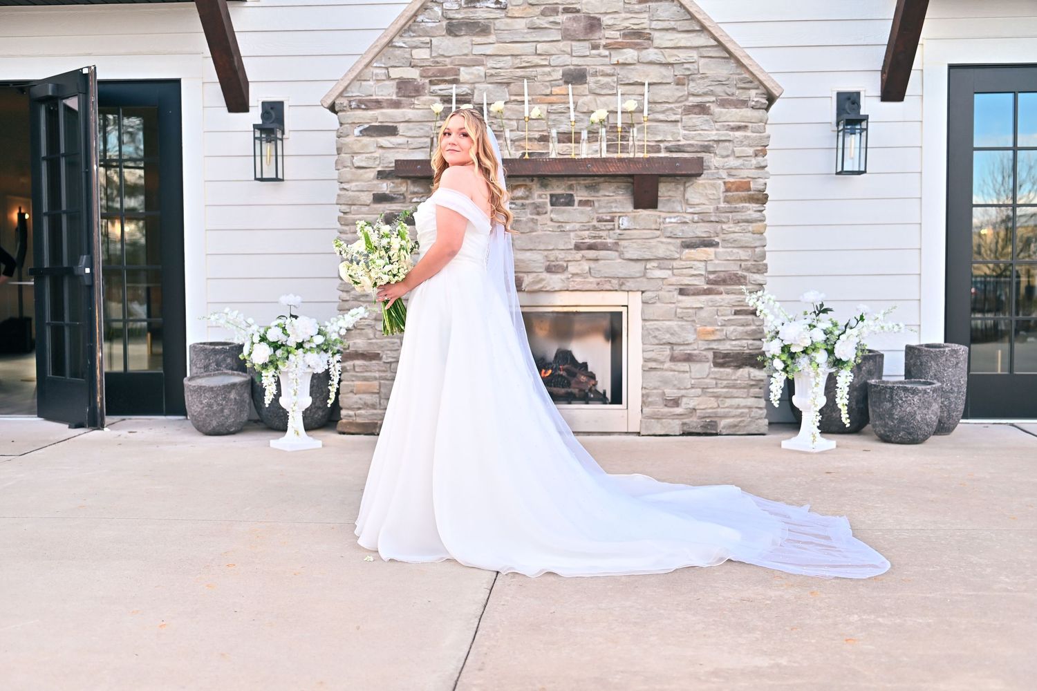 Bride in white off-shoulder gown with long train holds bouquet before stone fireplace at elegant outdoor venue.
