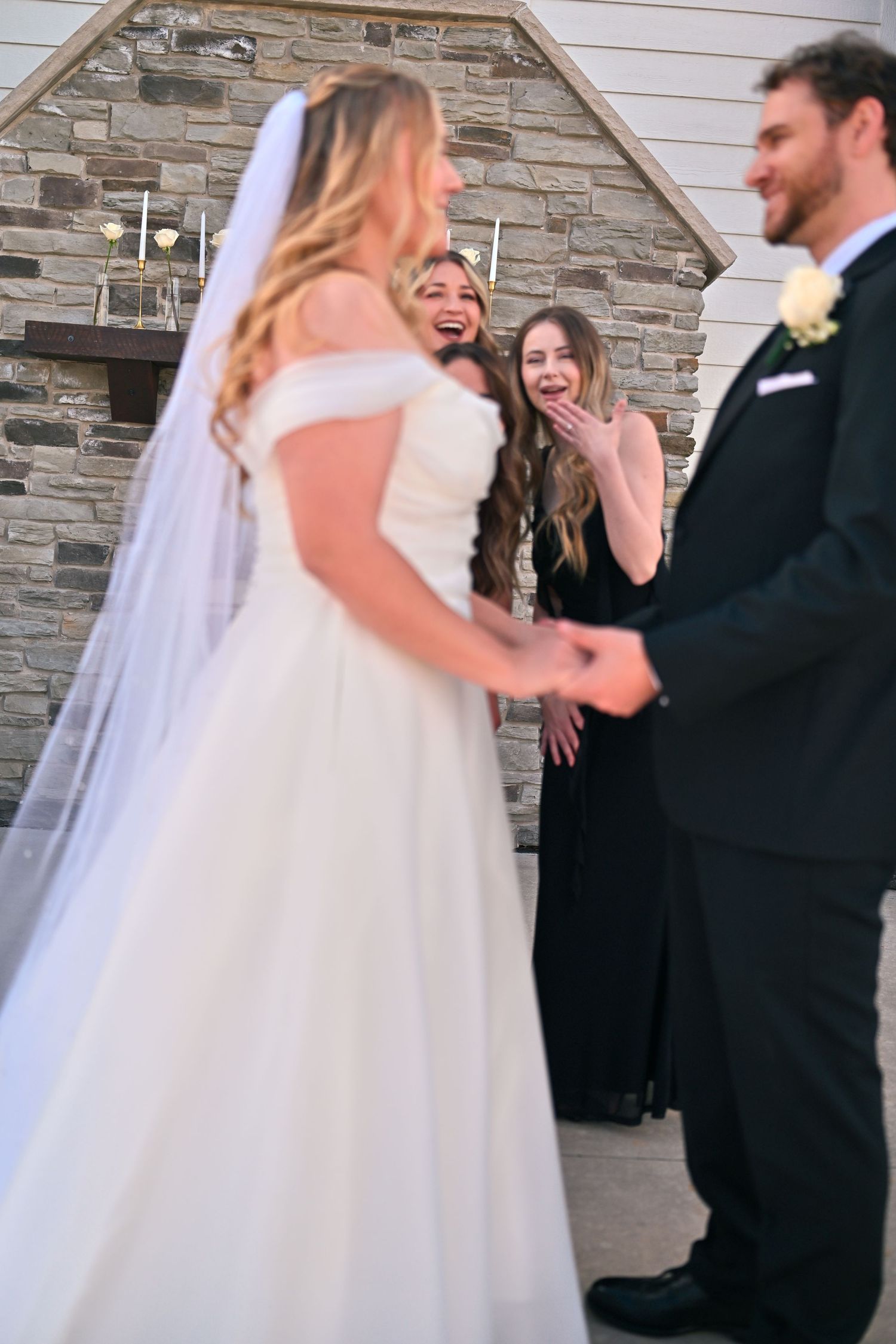 Bride in white gown and veil holds hands with groom in tuxedo as bridesmaids in black dresses react with joy.