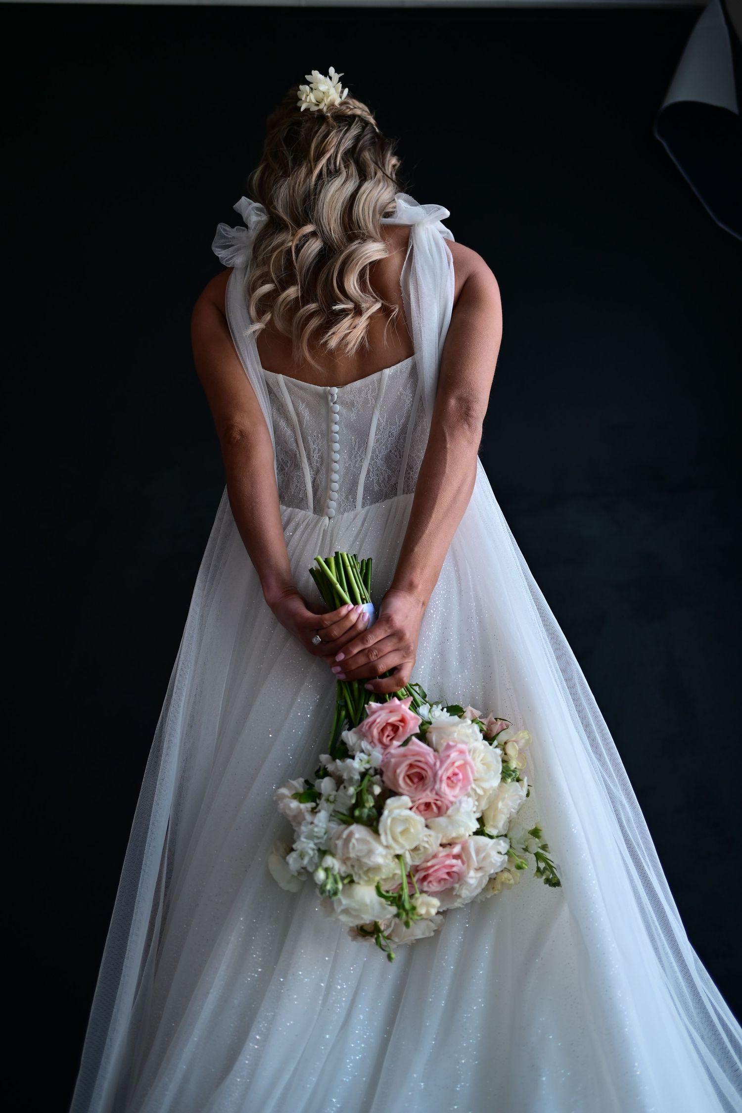 A bride in a flowing white wedding gown holds a romantic bouquet of pink and white flowers behind her back.
