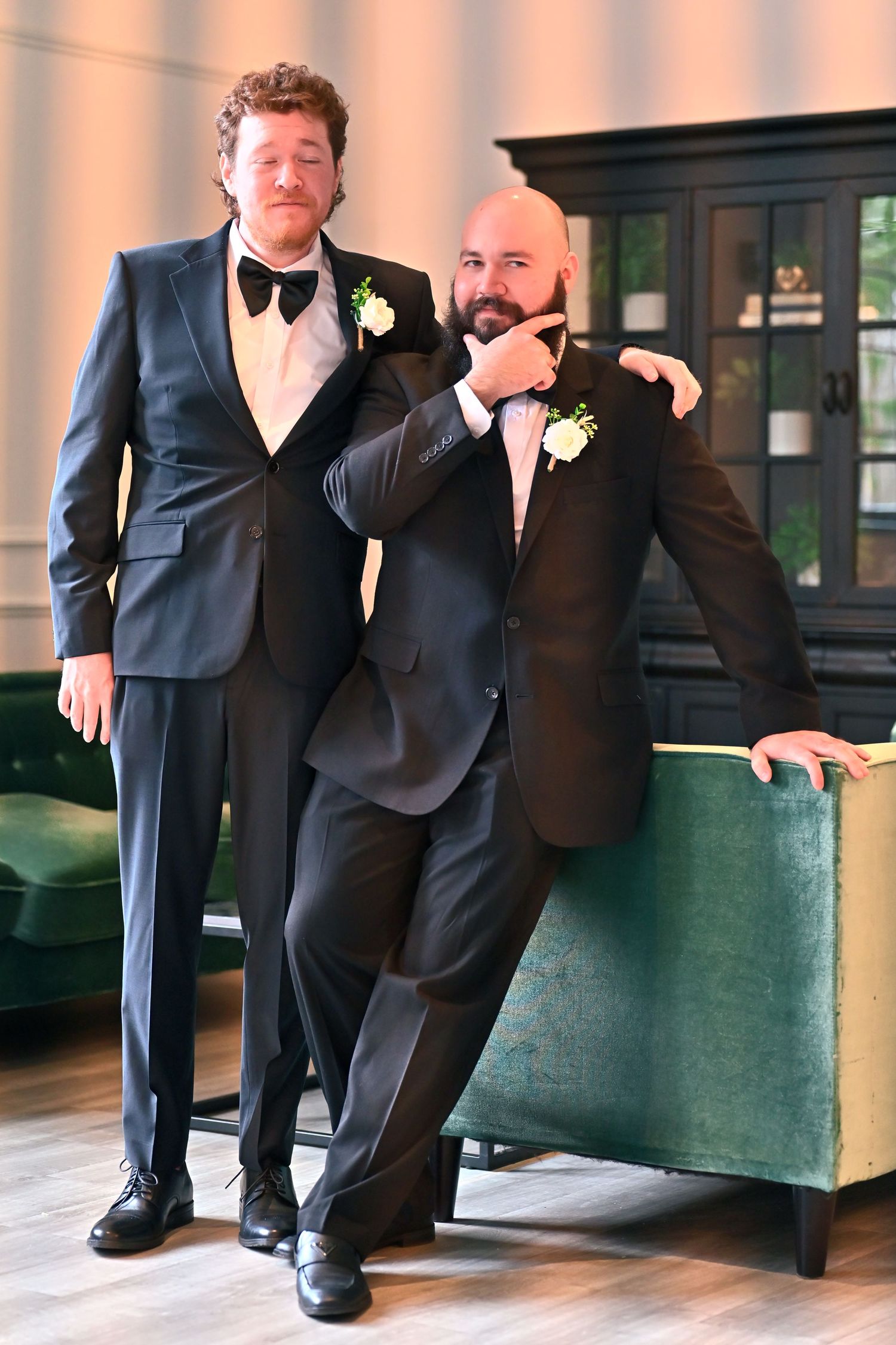Two grooms in black tuxedos with boutonnieres pose together in an elegant venue on their wedding day.