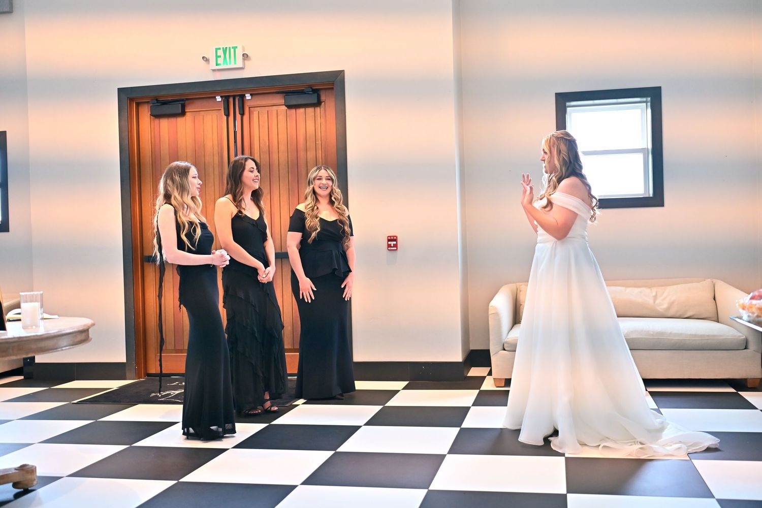 Bride in white gown shares emotional first look moment with bridesmaids in black dresses on checkered floor.