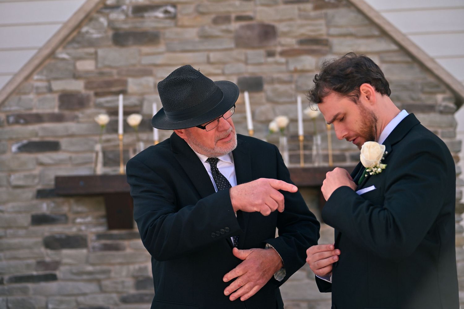Two men in formal attire adjust boutonnieres outside a stone church at a wedding ceremony.