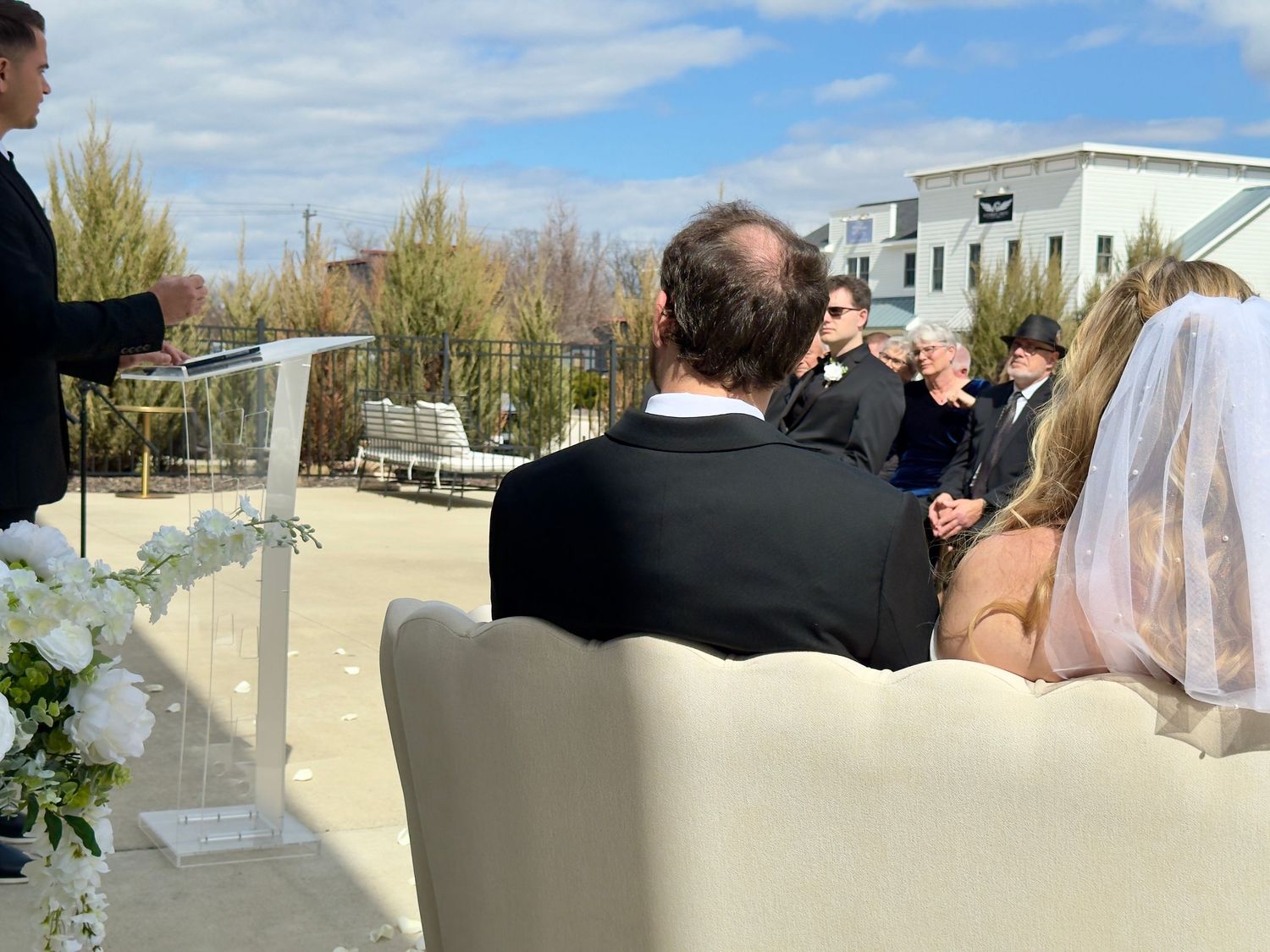 Outdoor rooftop wedding ceremony with bride in veil and groom seated on white sofa while officiant speaks at podium.