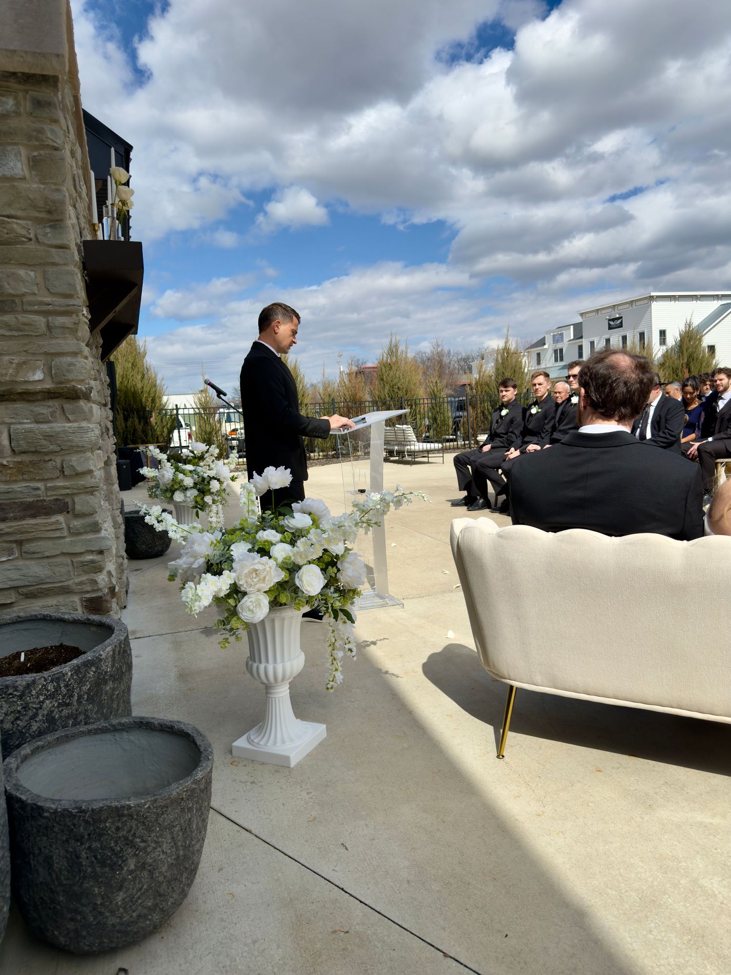 Outdoor wedding ceremony with officiant at podium, white floral arrangements, and guests seated on elegant furniture under cloudy sky.