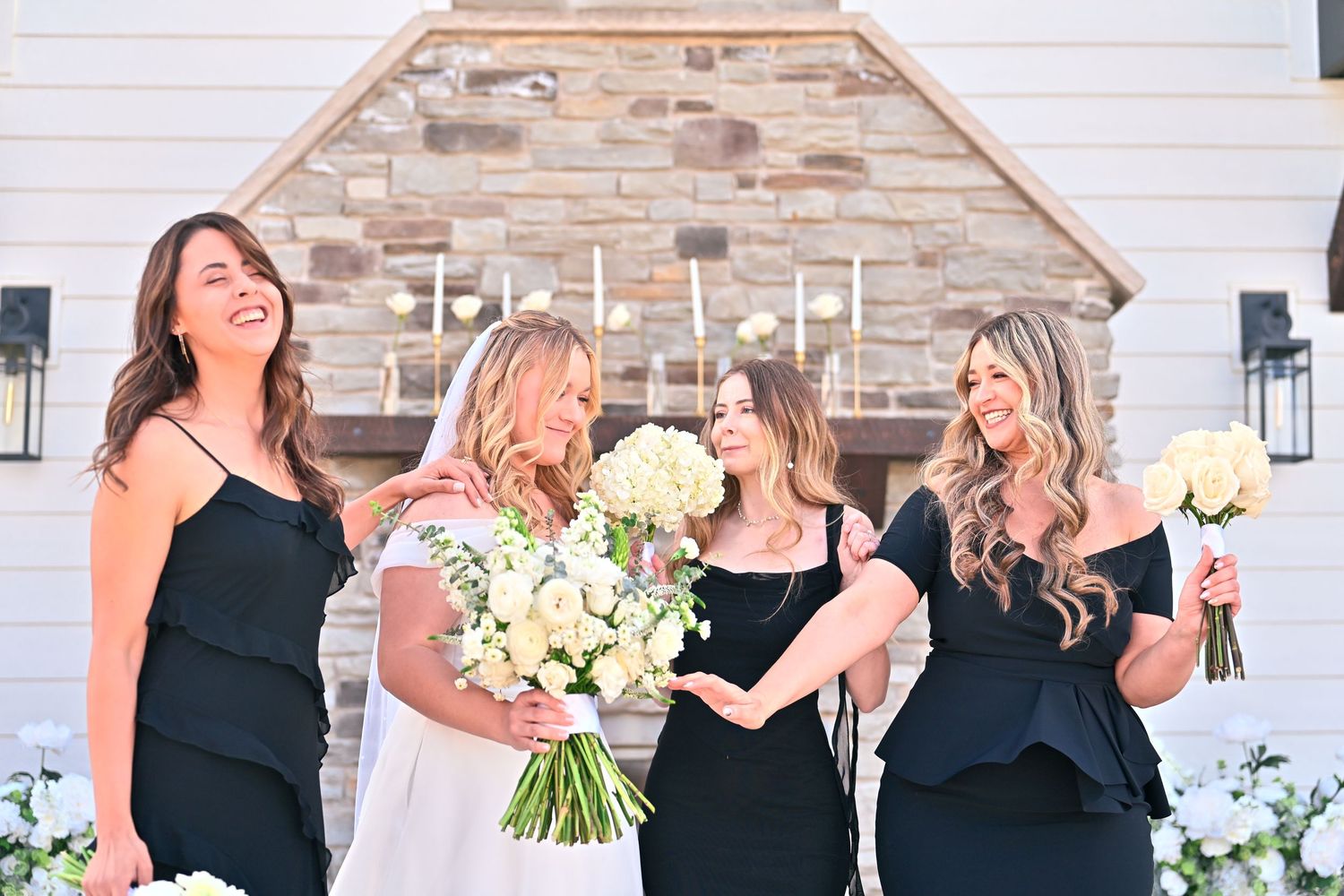 Bride in white gown with three bridesmaids in black dresses laughing outdoors holding white bouquets.