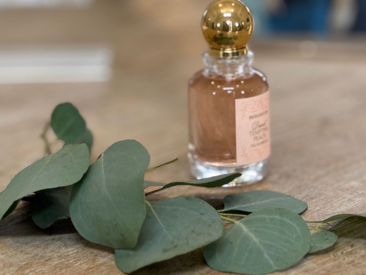 Glass perfume bottle with gold cap beside eucalyptus leaves on wooden surface.