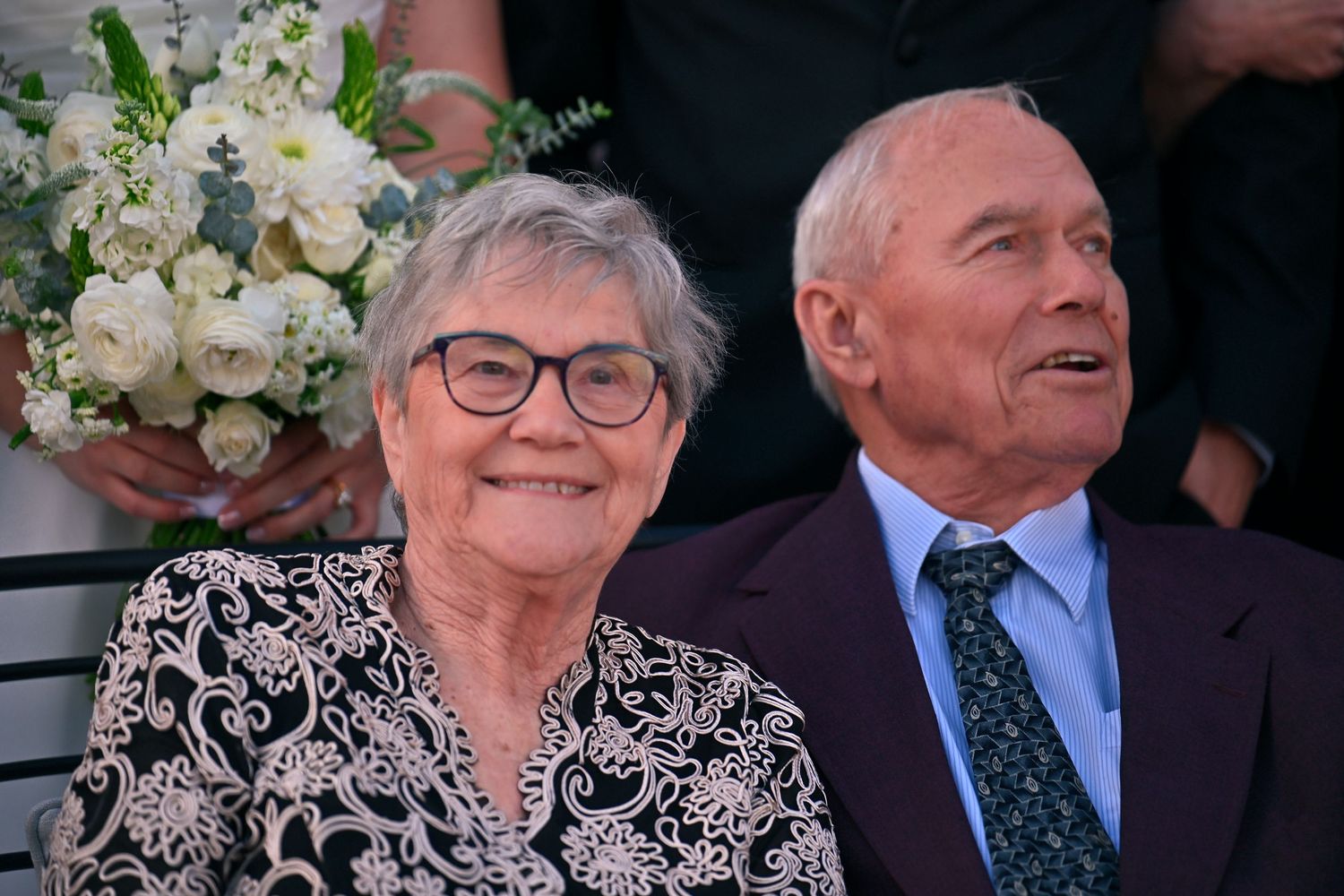 Elderly couple smiling at outdoor event, woman in floral blouse, man in suit with tie, white flowers in background.