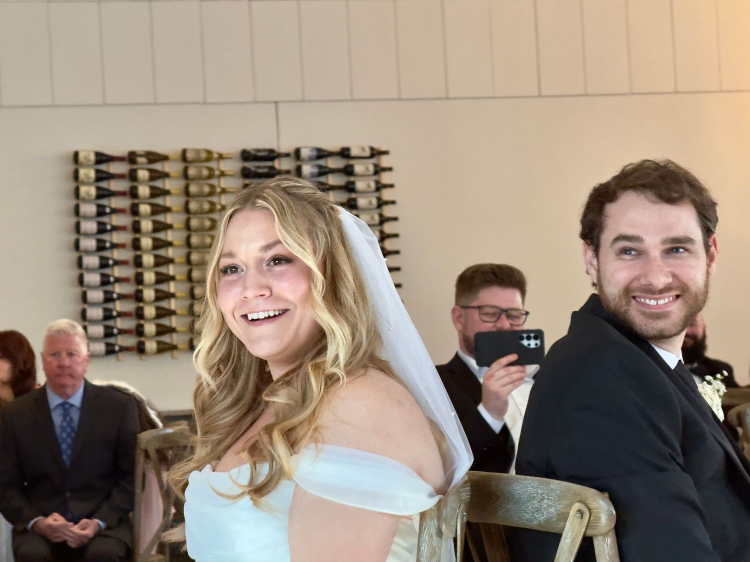 Smiling bride in white off-shoulder gown and veil seated with groom in tuxedo during indoor wedding ceremony.