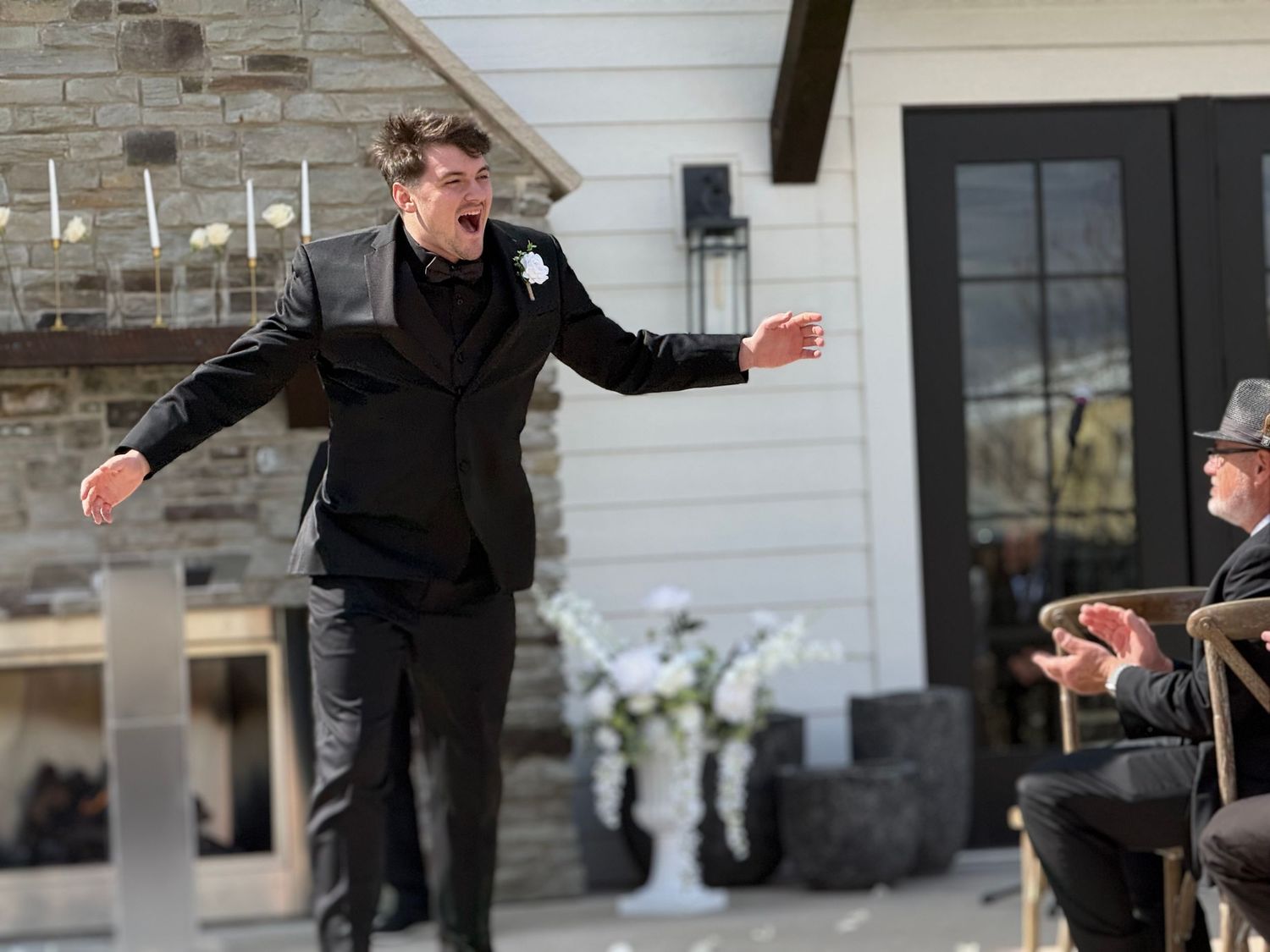 Man in black suit gesturing with outstretched arms outdoors at a wedding ceremony near a modern home.
