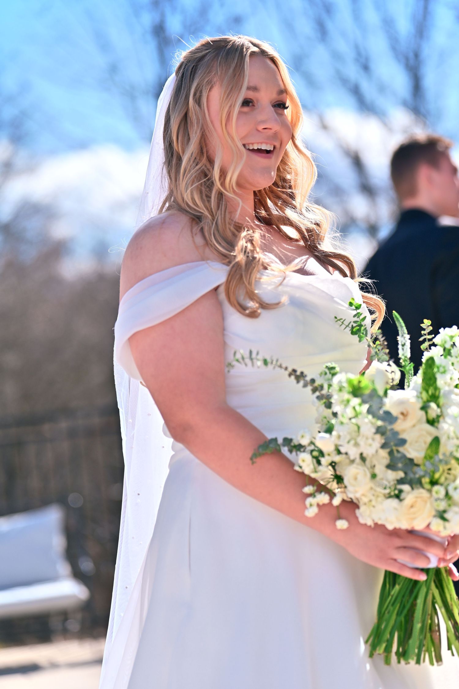 Smiling bride in white off-shoulder gown holding white floral bouquet outdoors on sunny day.