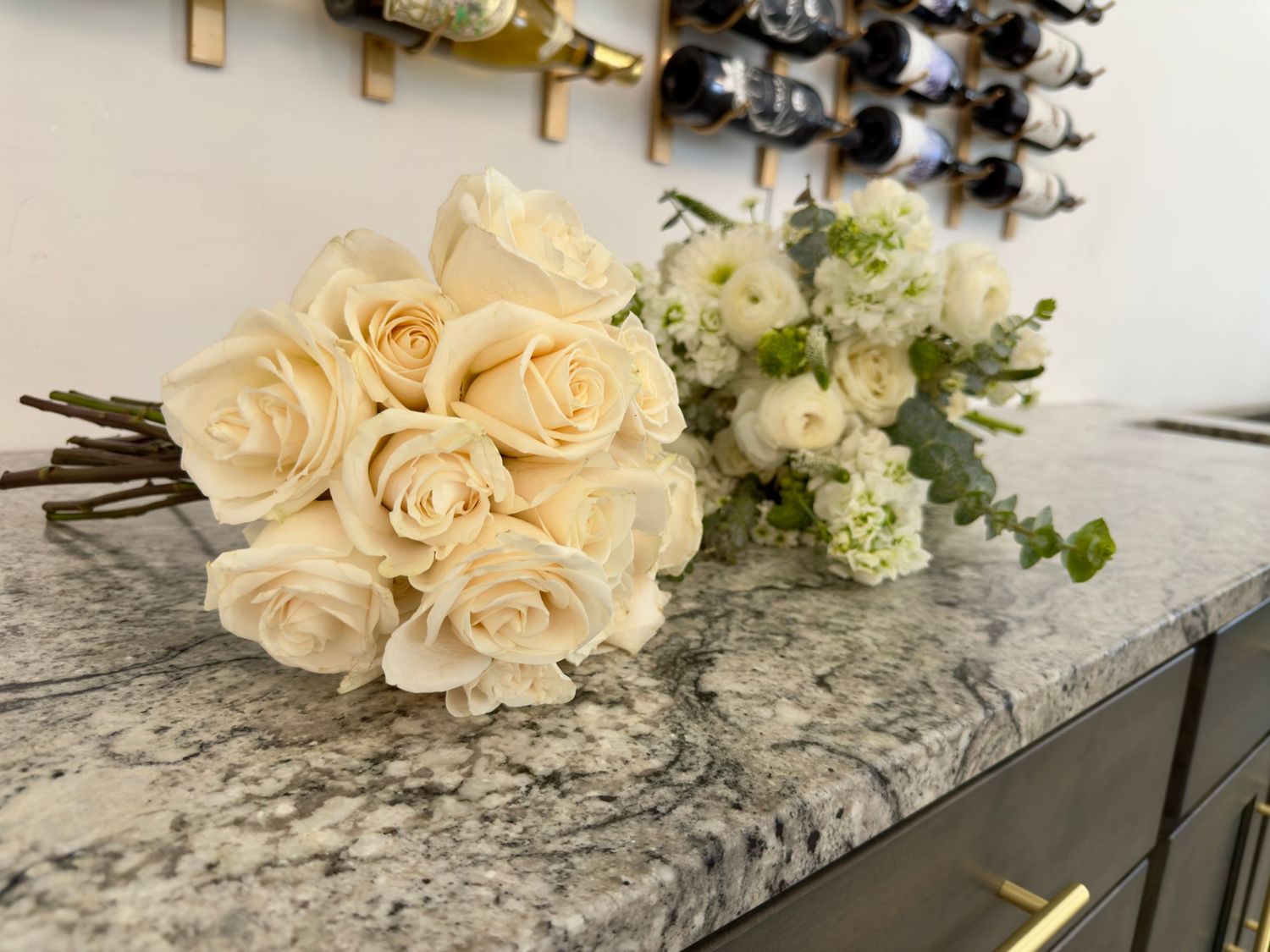 Two elegant white rose bouquets rest on a gray marble countertop near a wine rack.