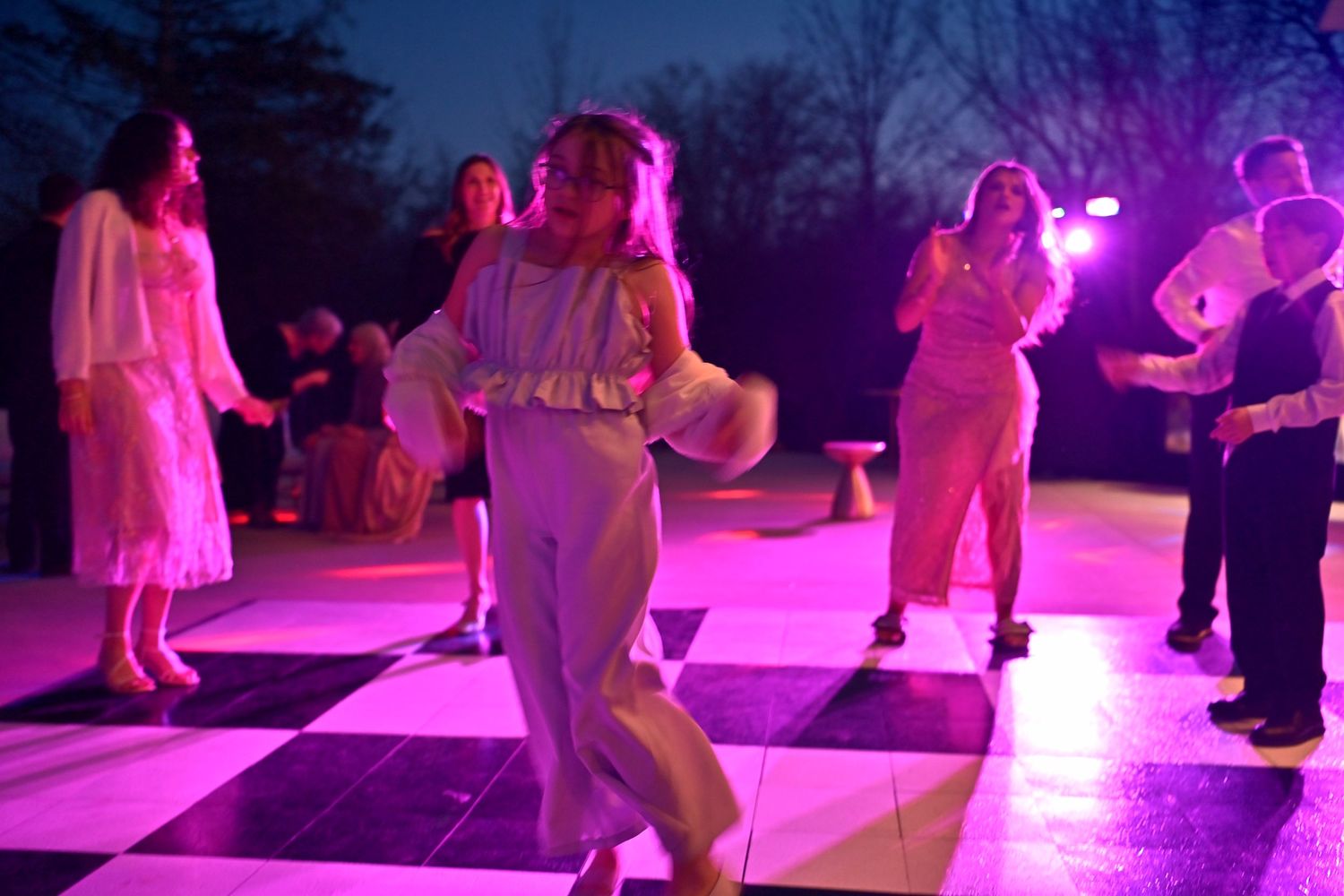 People dancing on a black and white checkered dance floor at an outdoor evening event with pink lighting.