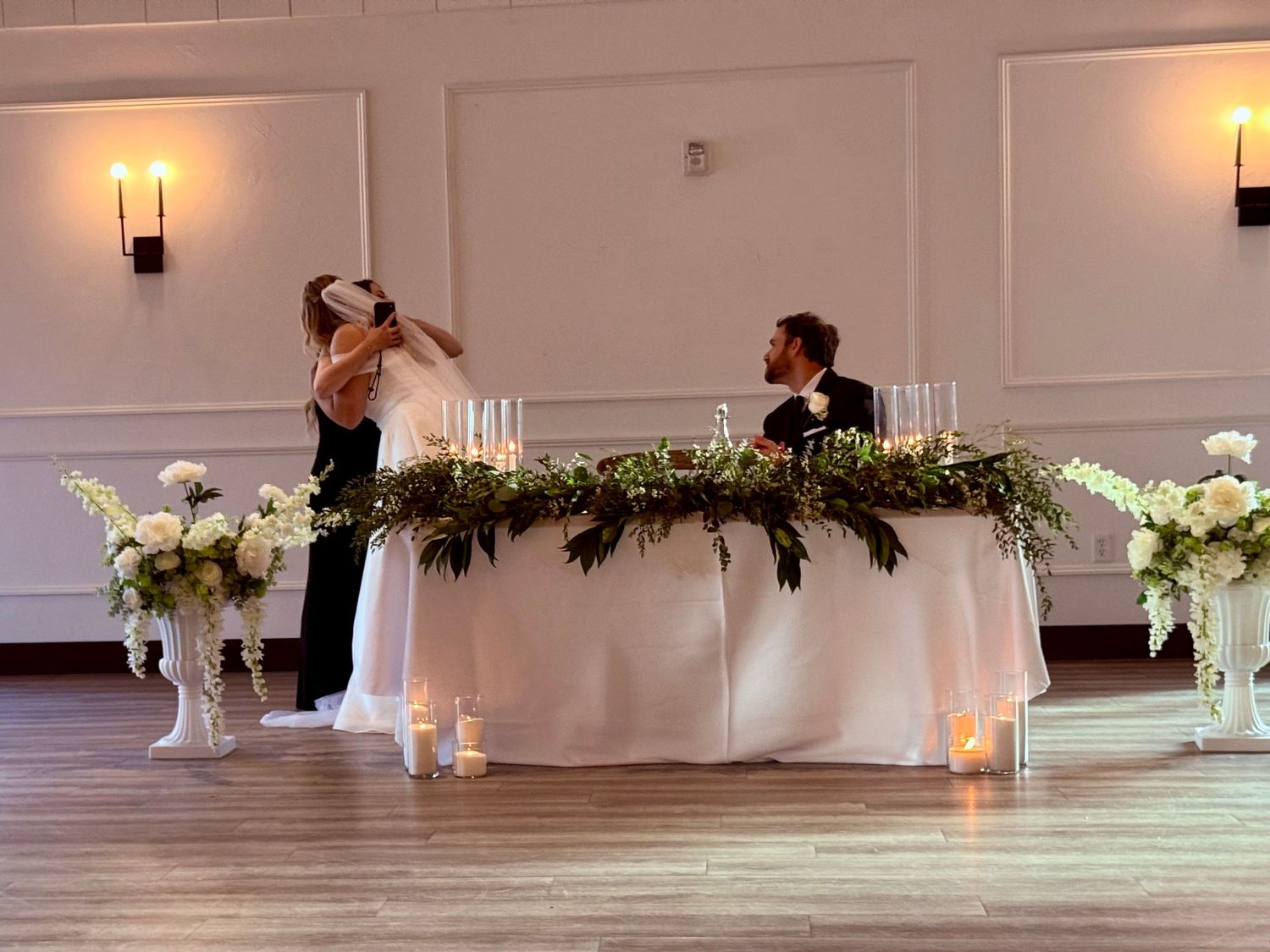 Elegant wedding reception sweetheart table with greenery garland, white florals, and candles in a bright ballroom.