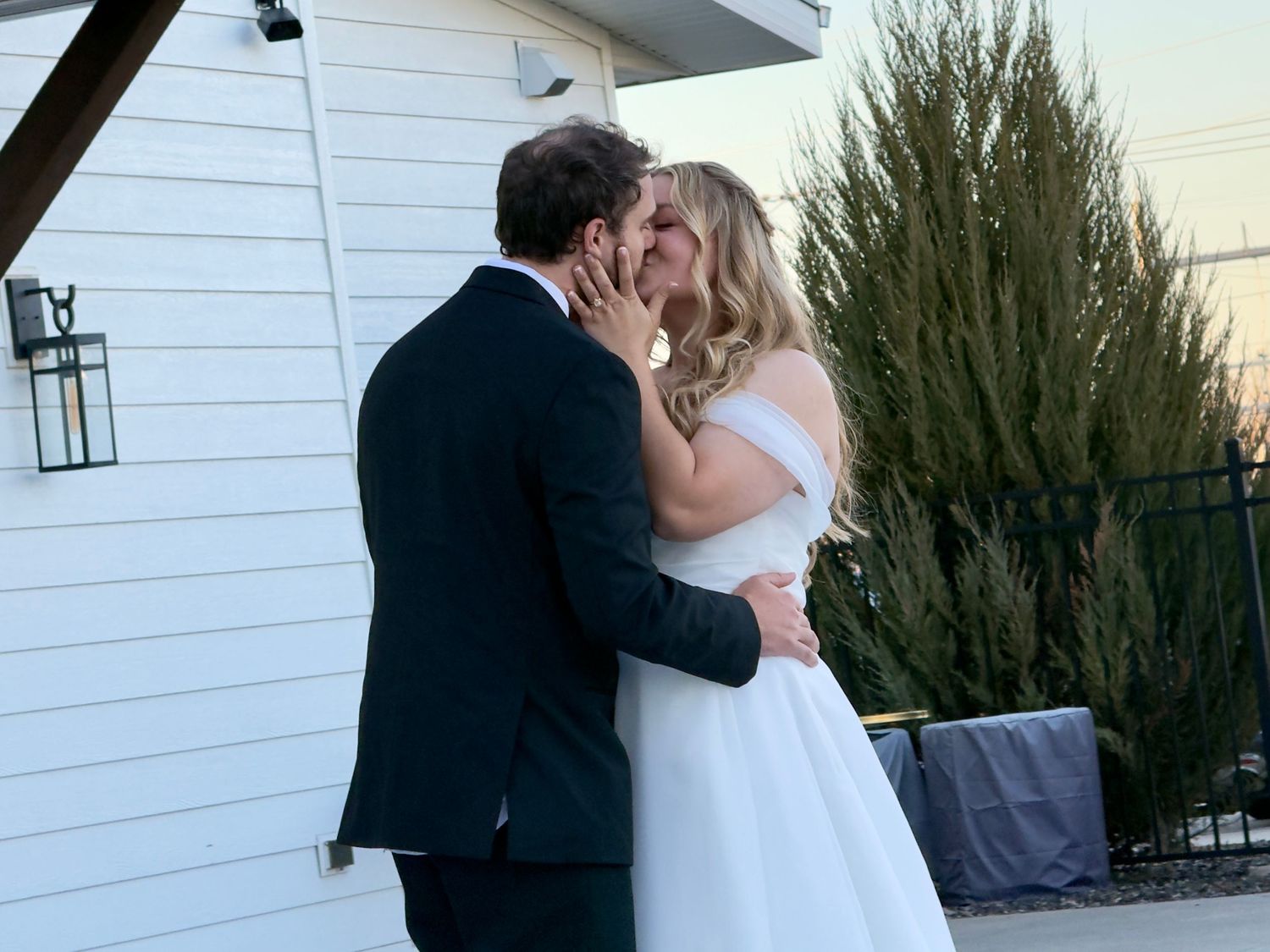 Bride and groom share a romantic first kiss outside a white venue at dusk on their wedding day.